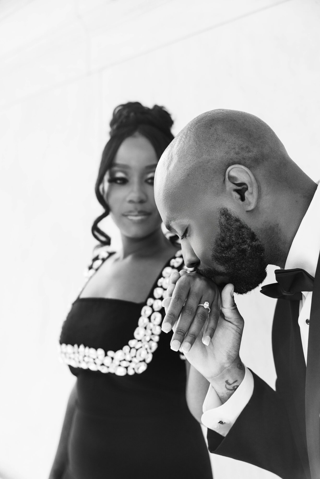 Man in tuxedo with bow tie and woman in dress with pearl embellishments, holding hands, in black and white.