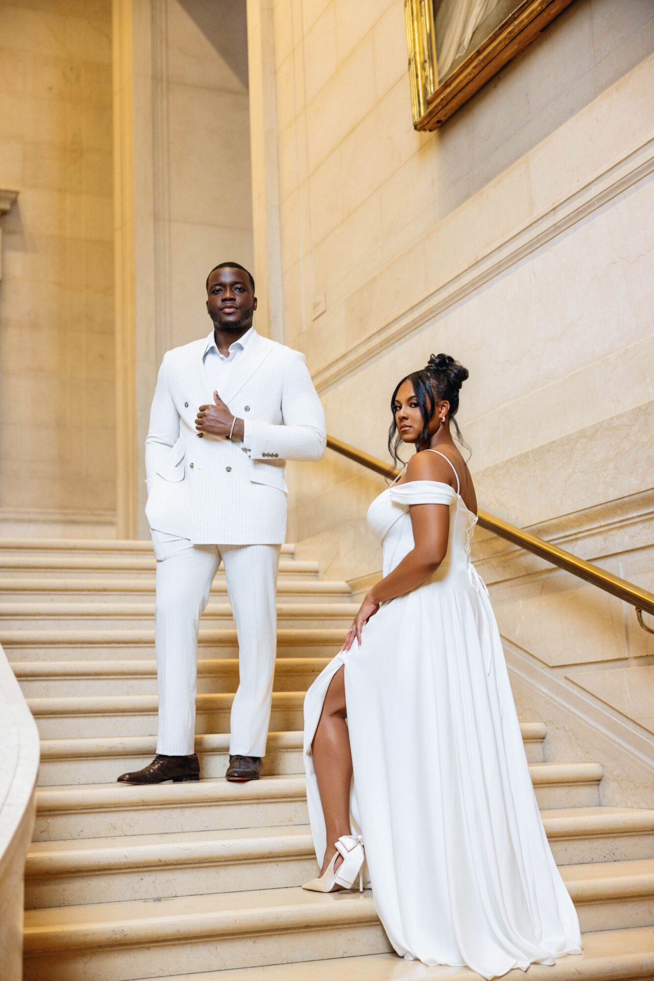 A man and woman in white formal attire standing on a staircase inside a building with beige walls and a gold-framed mirror.