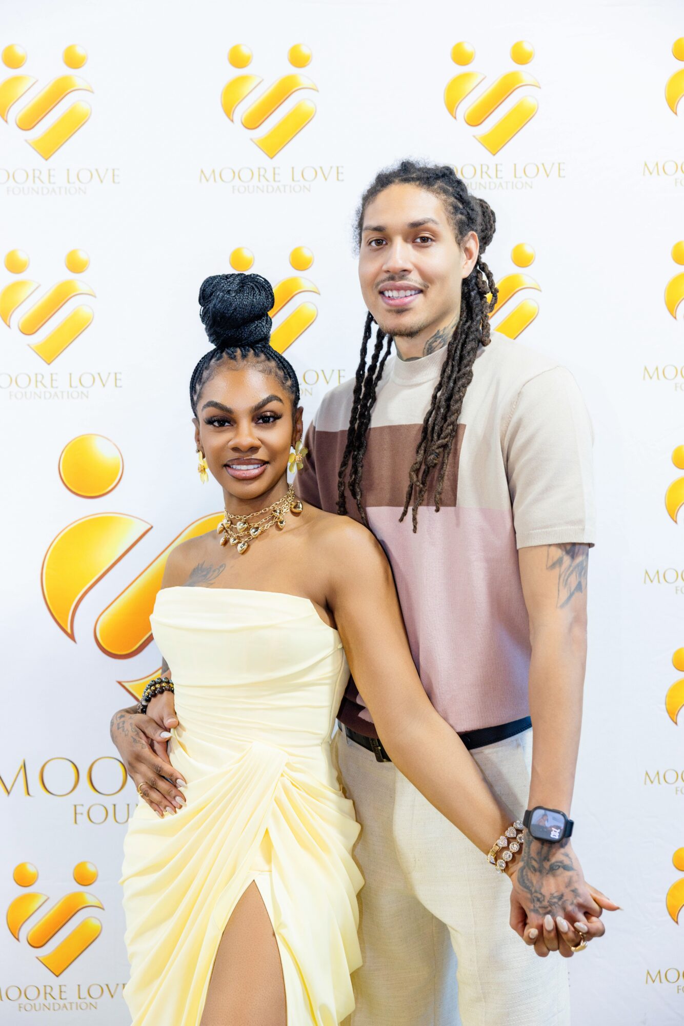 Two people smiling, standing in front of a white background with yellow logo and text, holding hands.