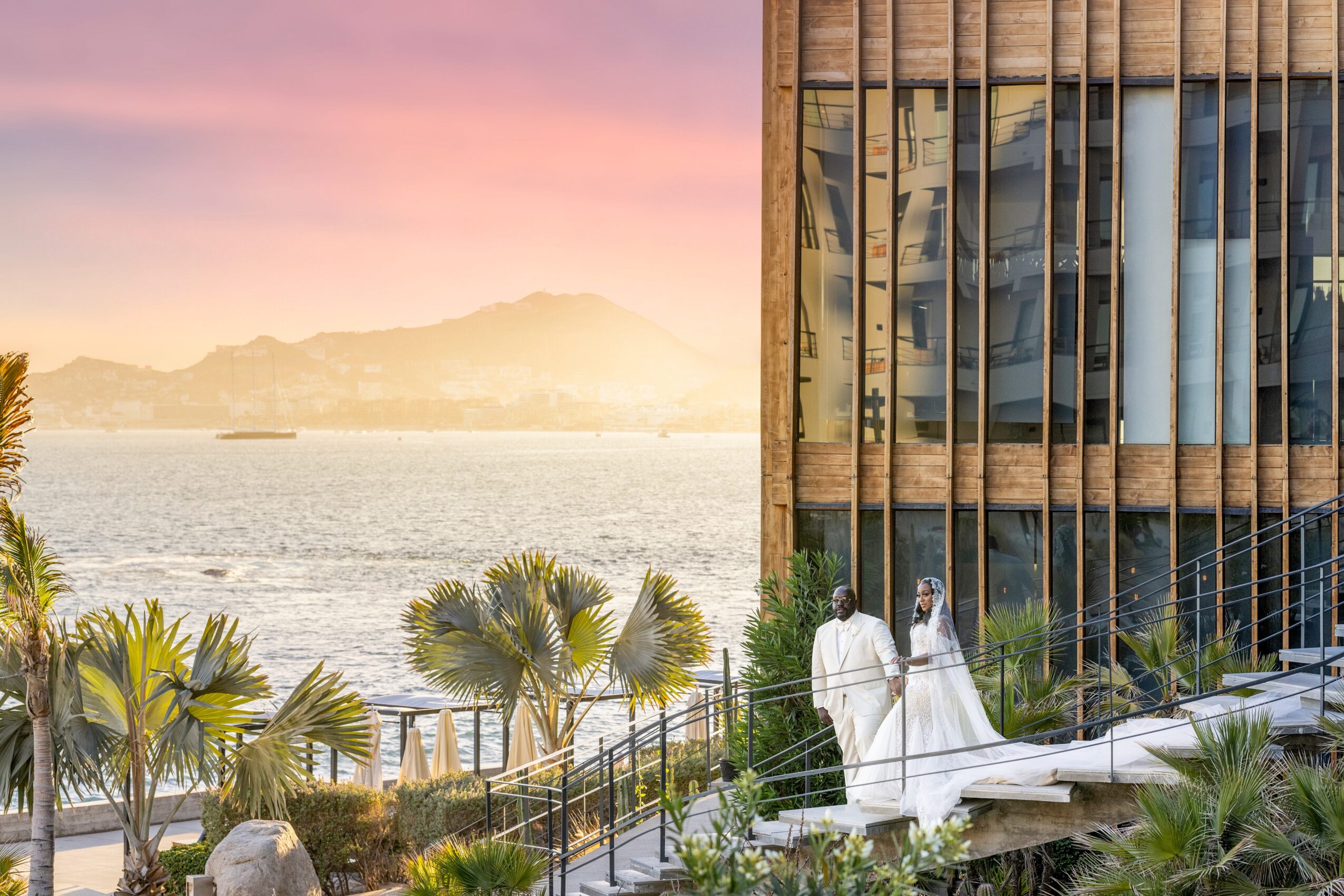 Two people in white clothing walk along a pathway near a modern glass building with plants and water in the background.
