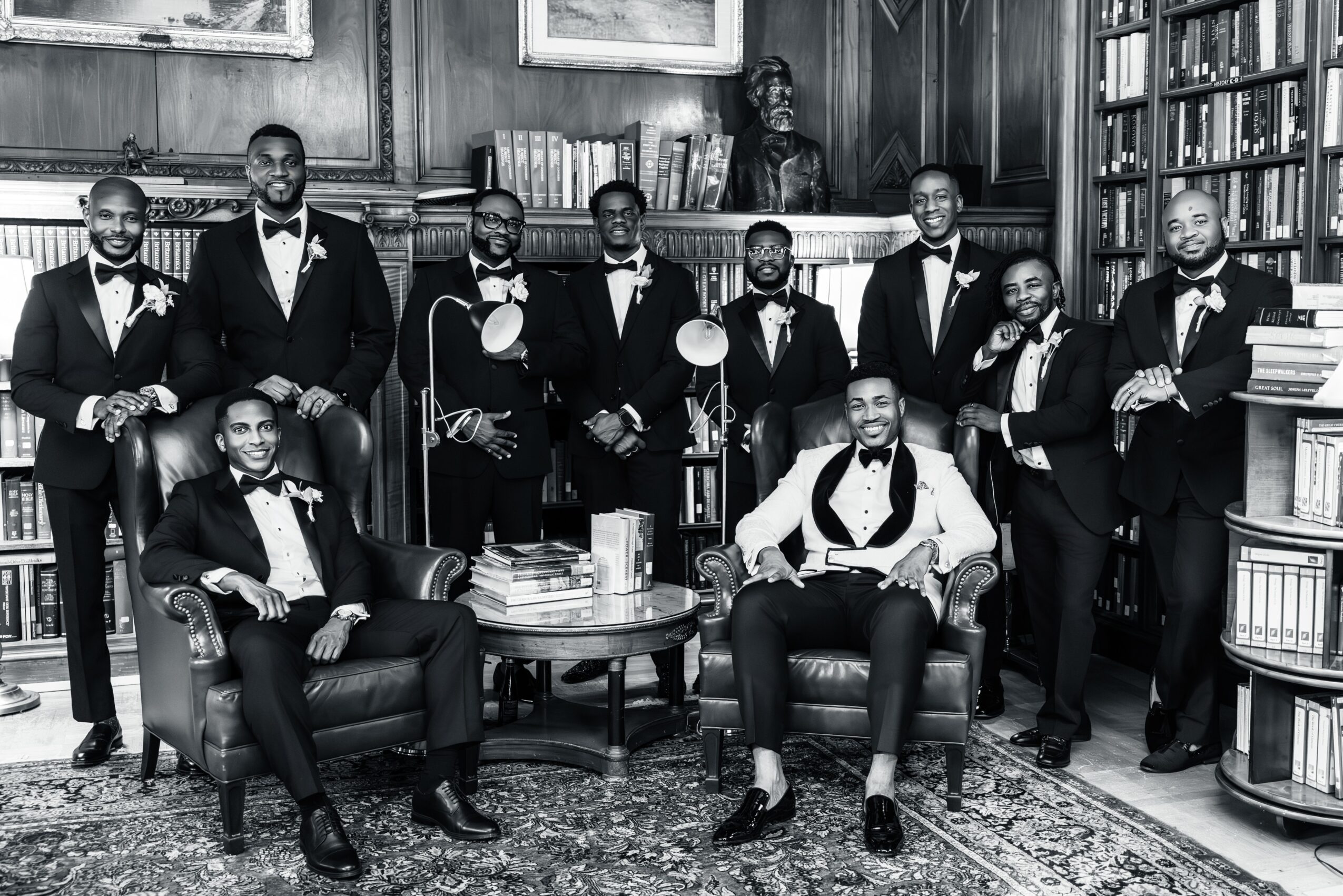 Group of men in tuxedos in a library room with bookshelves and a wooden desk, some seated and others standing.