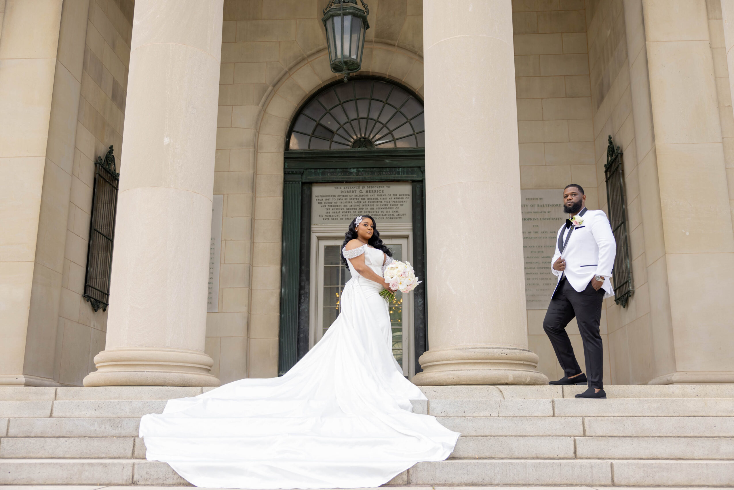Bride in a white wedding dress holding a bouquet, standing on steps between two large columns, with a groom in a tuxedo nearby.