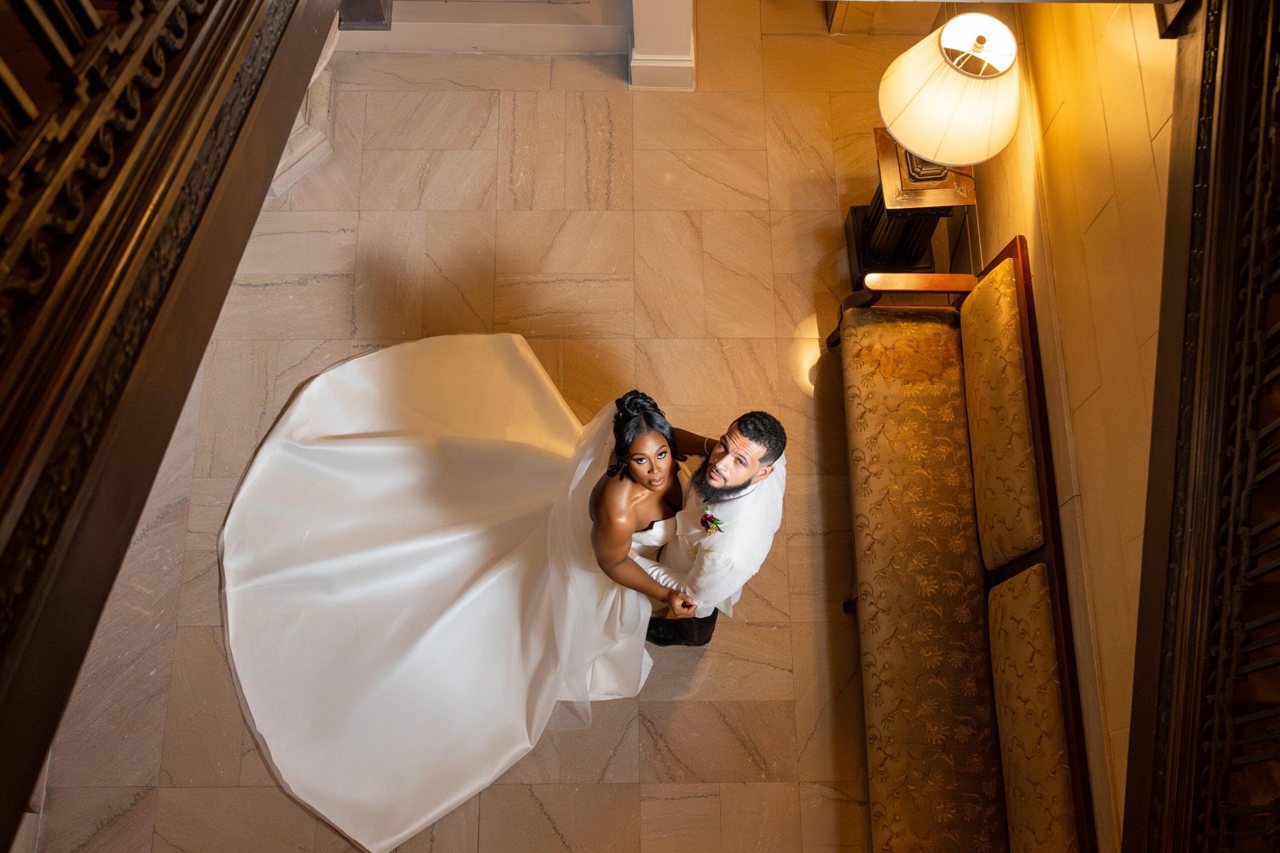 A bride in a white wedding dress and a groom in a suit stand together on a tiled floor, looking up at the camera.