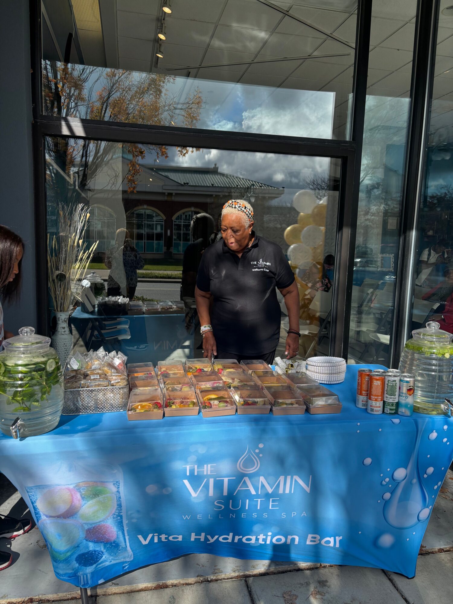 Person standing behind a table with snacks and drinks, in front of a glass window, with a blue banner reading 'VITAMIN Via Hydration Bar'.