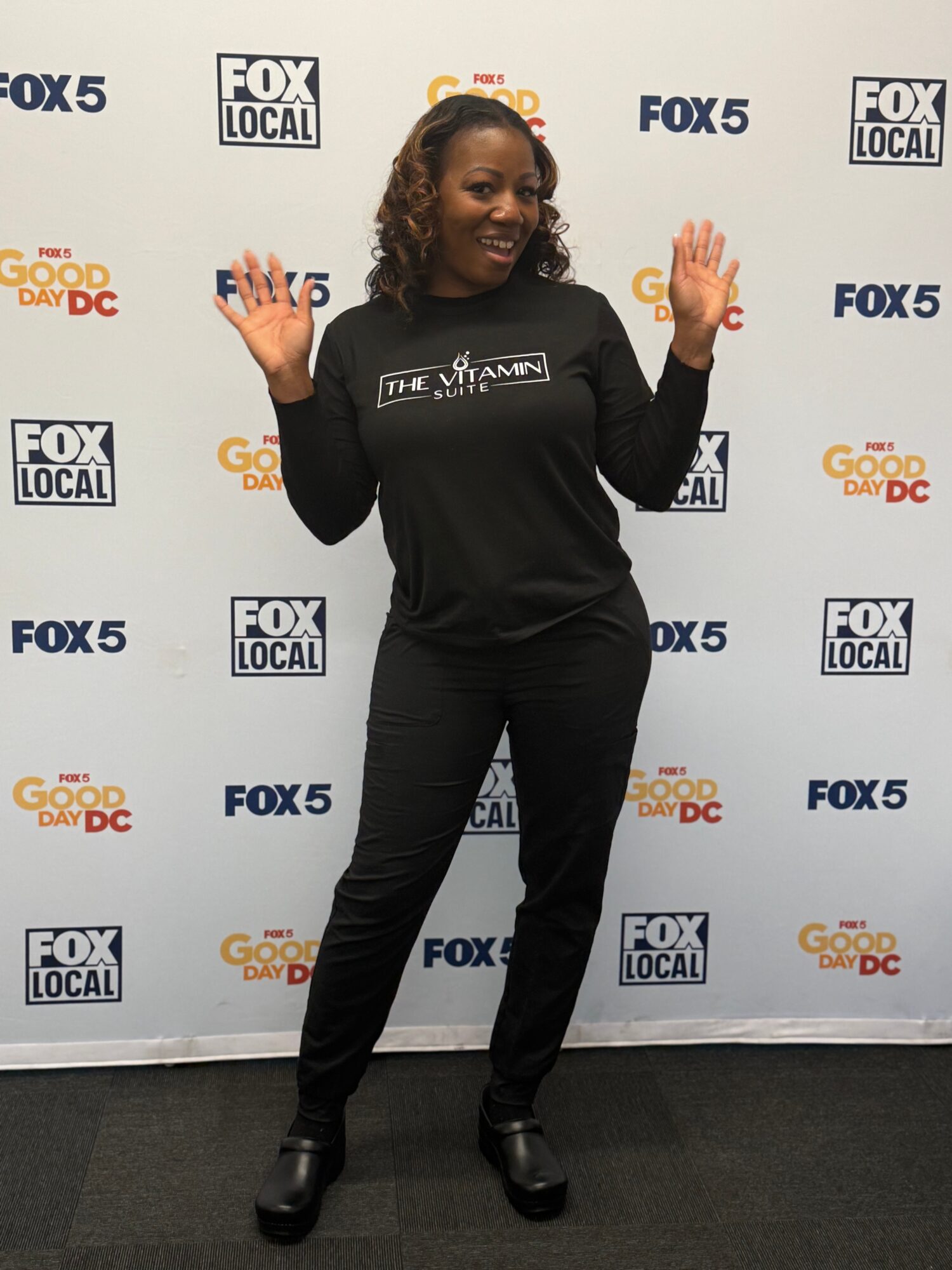 Woman standing with hands raised, smiling, in front of a backdrop with TV station logos, wearing a black outfit.