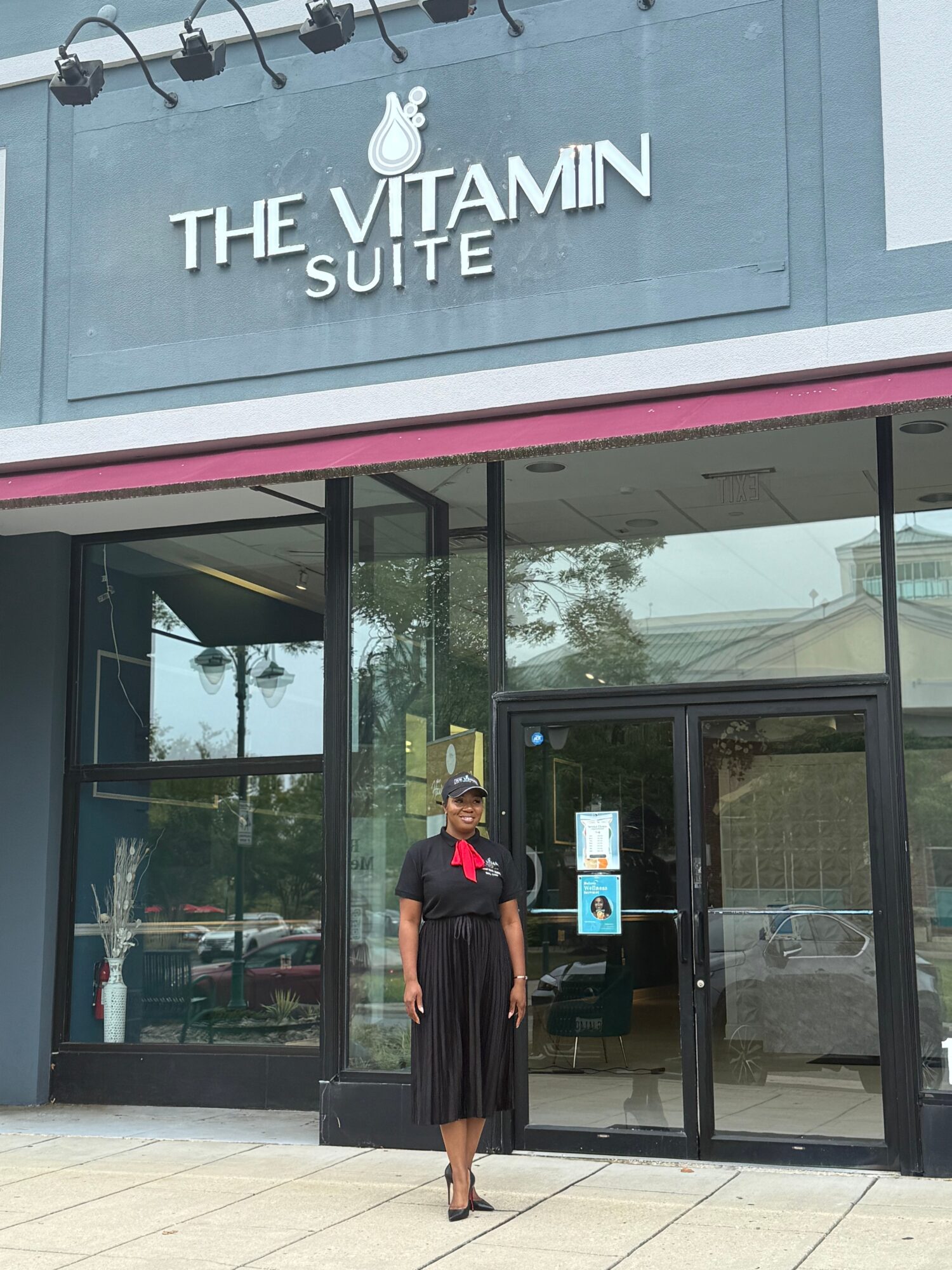 Person standing outside a glass storefront with sign 'The Vitamin Suite' above, wearing a black dress and sunglasses.