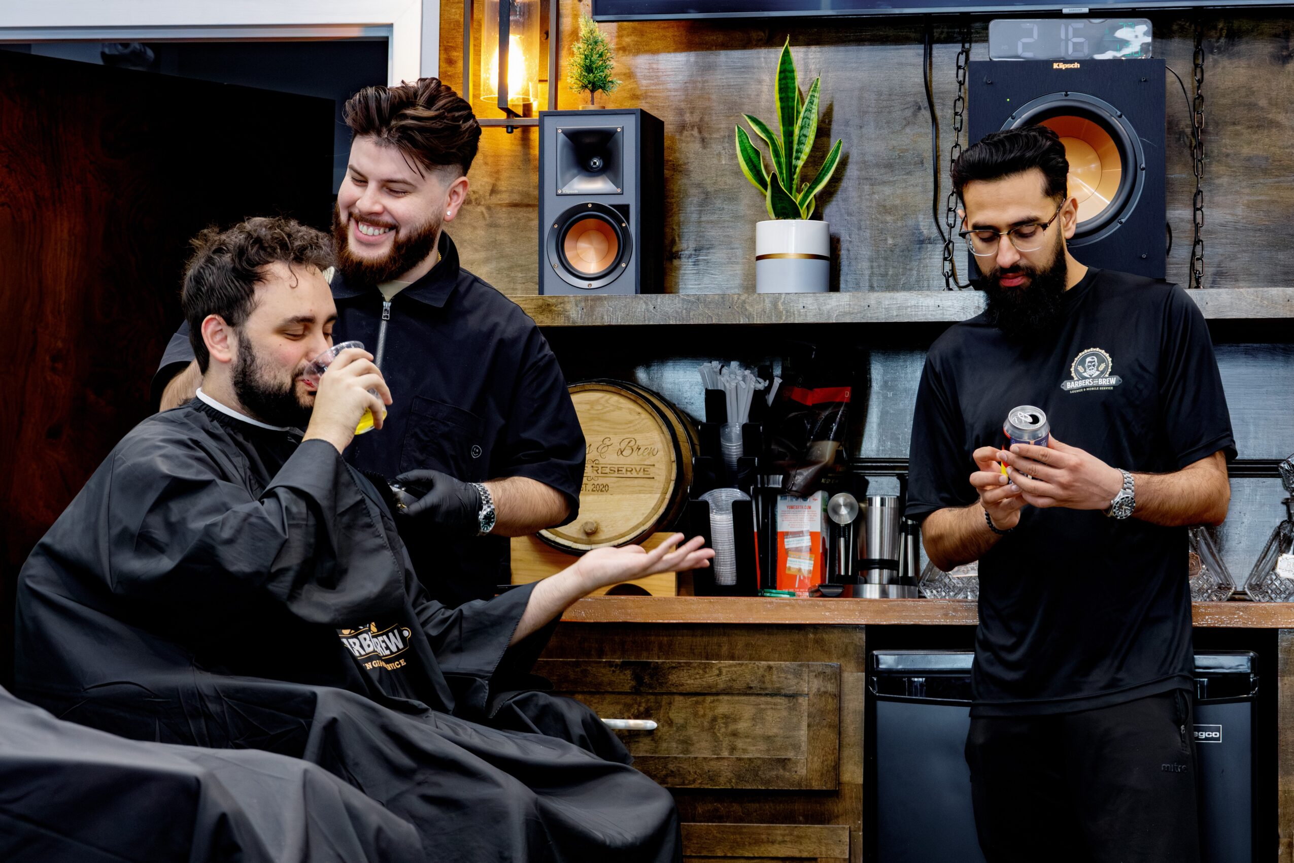Three men in a barbershop, one sitting in a barber chair, two standing, with shelves and decor in the background.