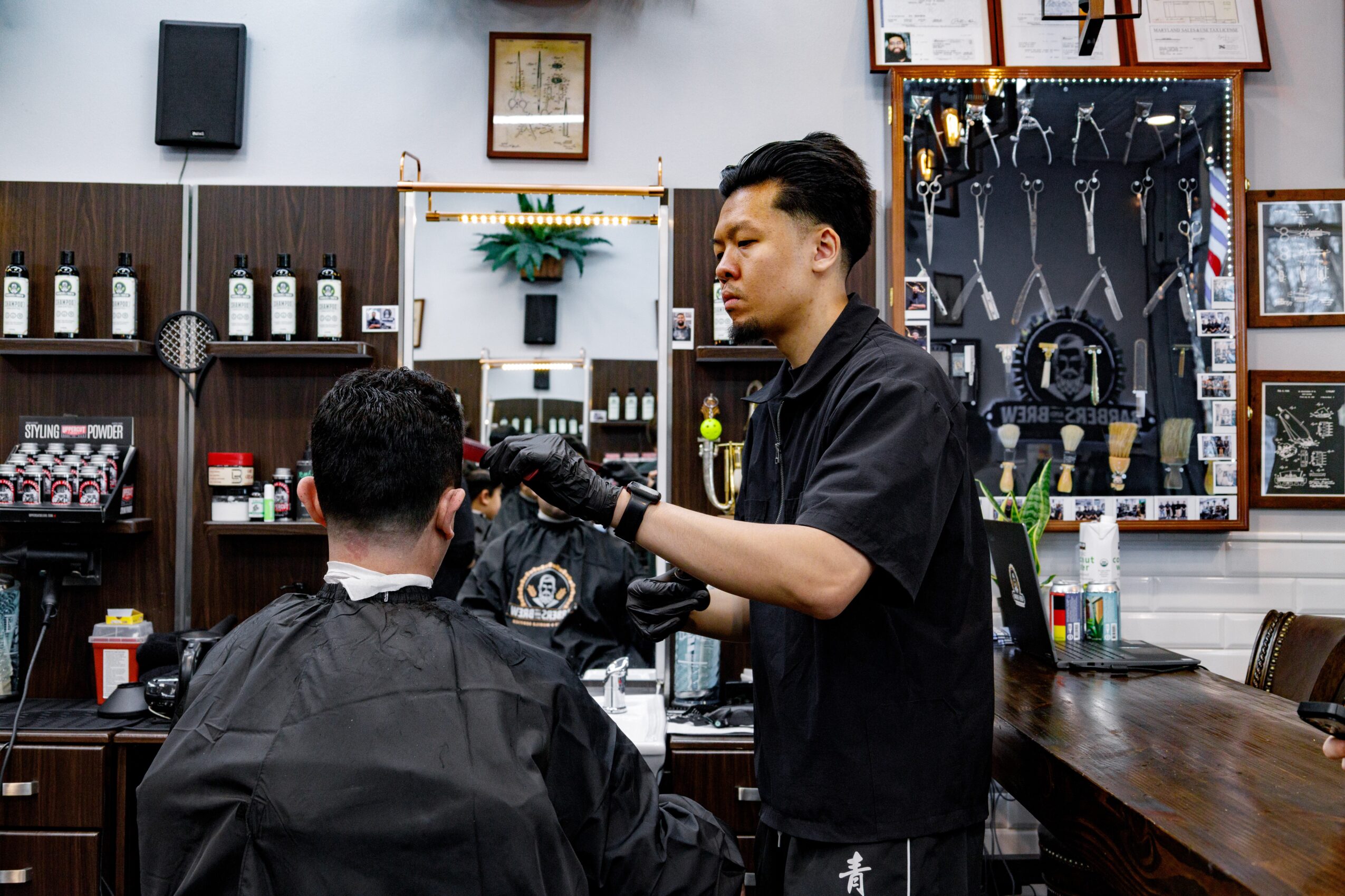 Barber cutting a customer's hair in a barbershop with tools and framed pictures on the wall.
