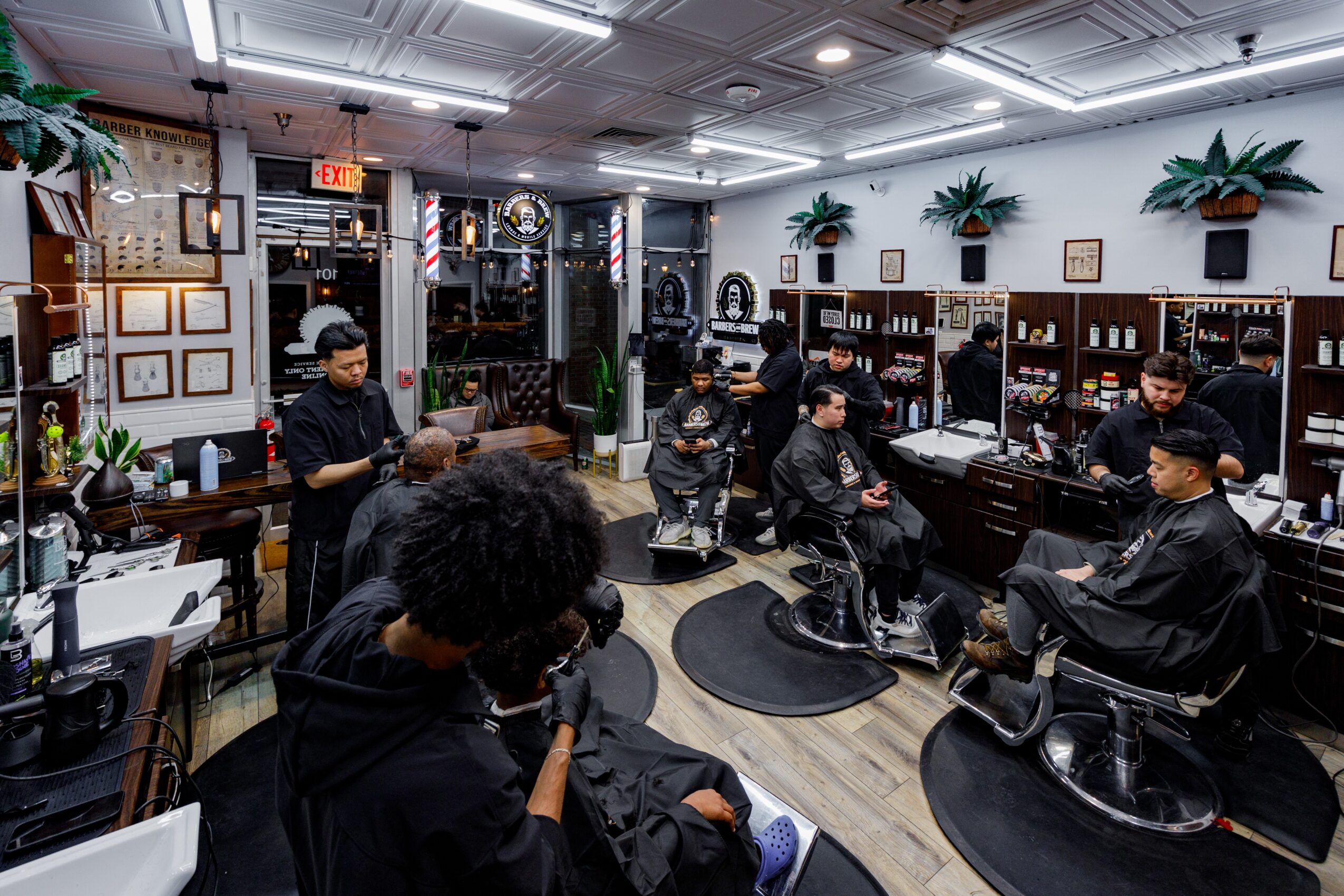 Barbershop interior with multiple barbers working on clients, shelves with products, and decorative plants on the wall.