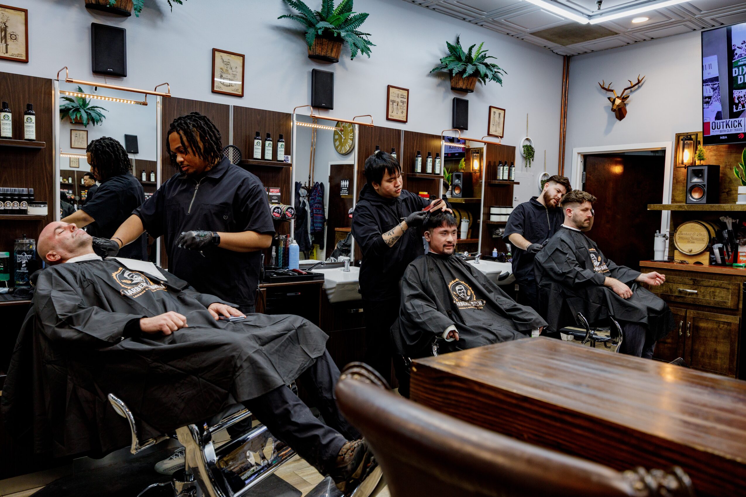 Barbershop with three clients getting haircuts, barbers working, and decor including plants, framed pictures, and a mounted deer head.