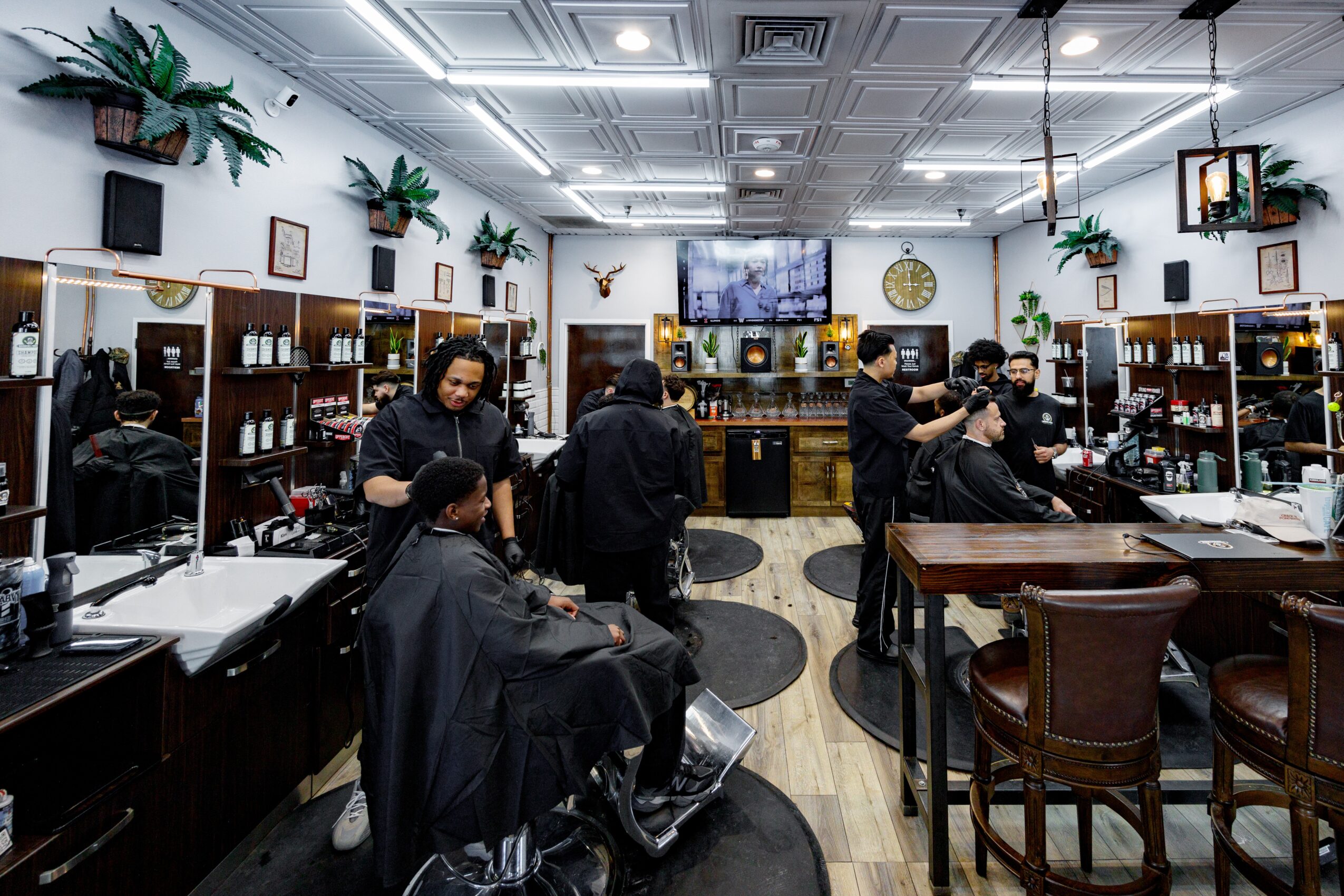 Barbershop interior with customers and barbers, mirrors, shelves, and a television, featuring wooden furniture and ceiling lights.
