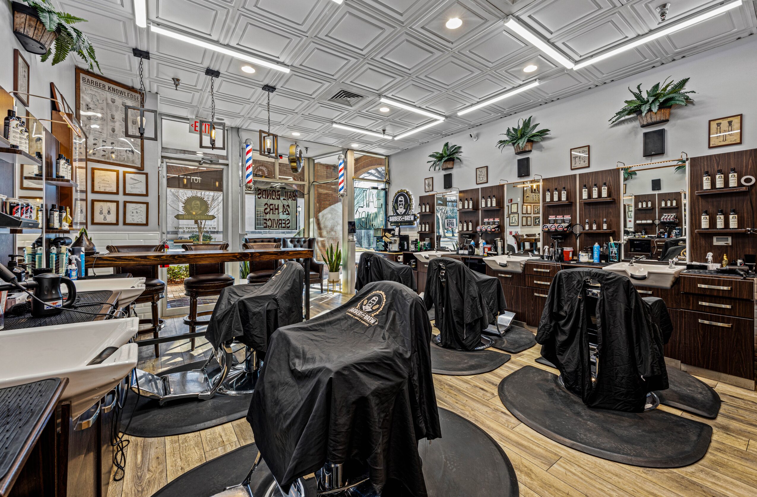 Barber shop interior with chairs covered in black capes, shelves with products, and large windows, decorated with plants and framed pictures.