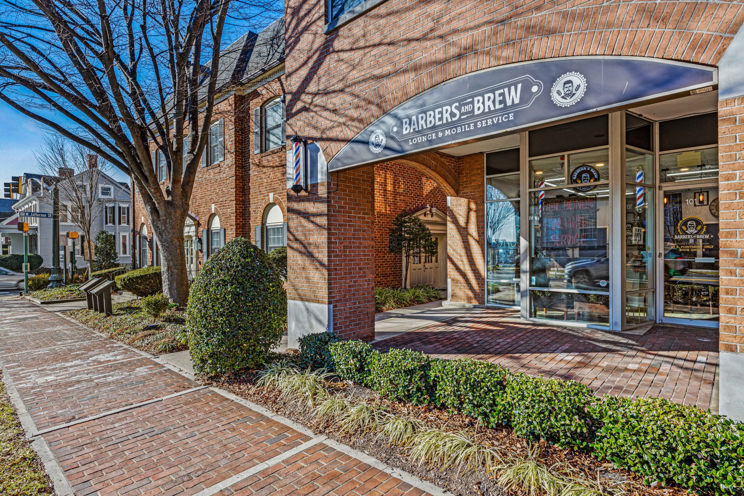 Street view of a building with a barbershop and brew shop, brick sidewalk, trees, and bushes, sunny day.