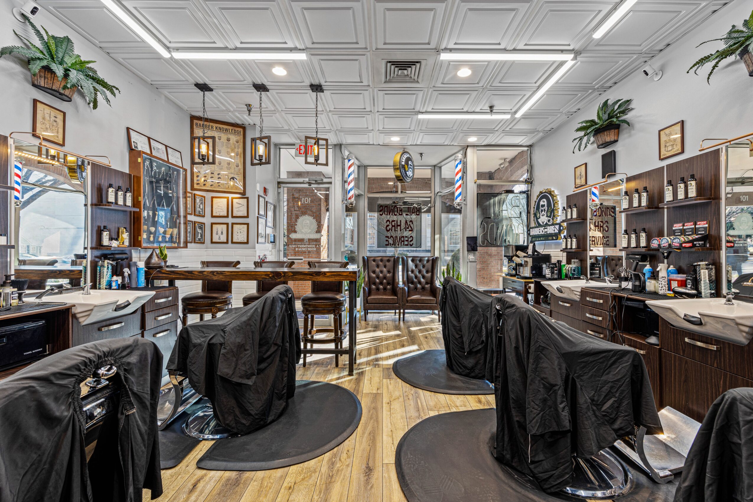 Barbershop interior with chairs, mirrors, shelves, and framed pictures, decorated with plants and ceiling lights, wooden flooring, and a ceiling with decorative molding.