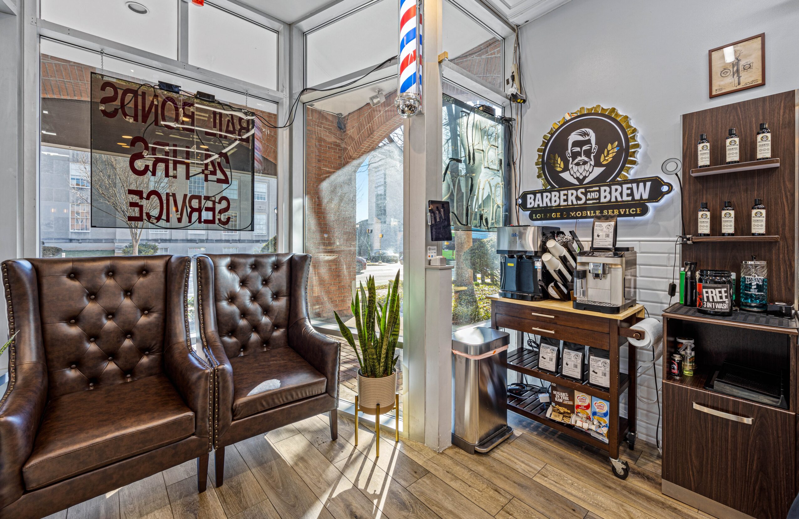 Barber shop interior with leather chairs, a potted plant, and barber supplies on shelves, seen through a window and door.