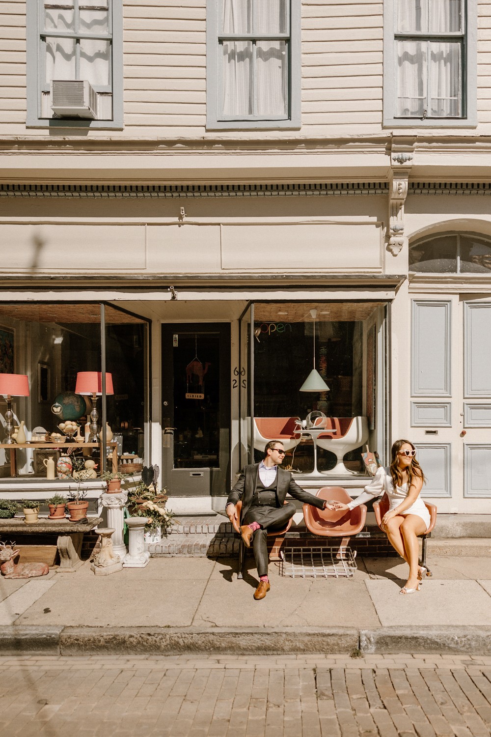 Two people sit on a bench outside a storefront with large windows and a door, in front of a building with white siding.
