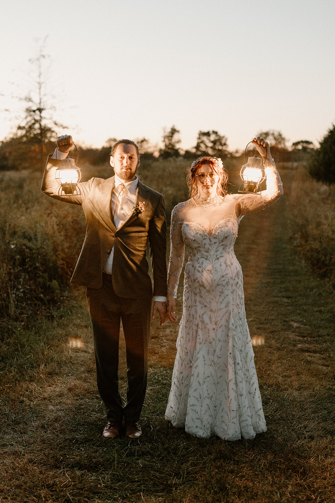 A bride and groom holding lanterns walk outdoors on a dirt path at sunset, with trees in the background.