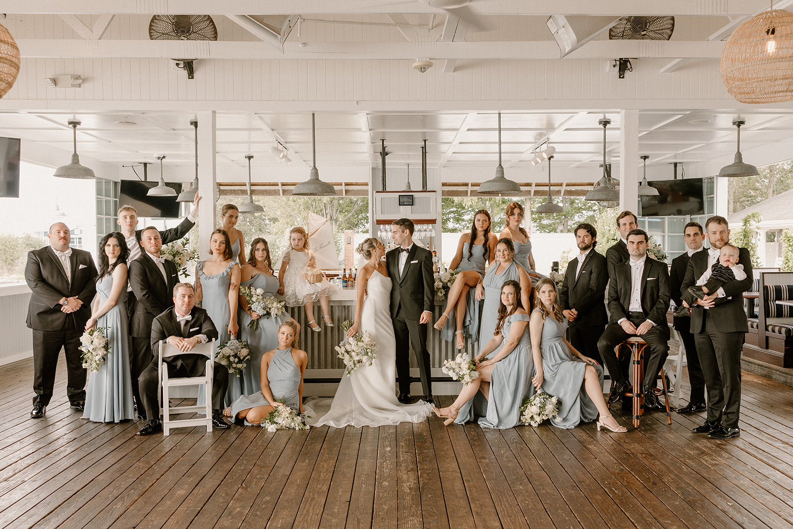 Group of people in formal attire posing indoors with a bride and groom in the center, wooden floor, and large windows.