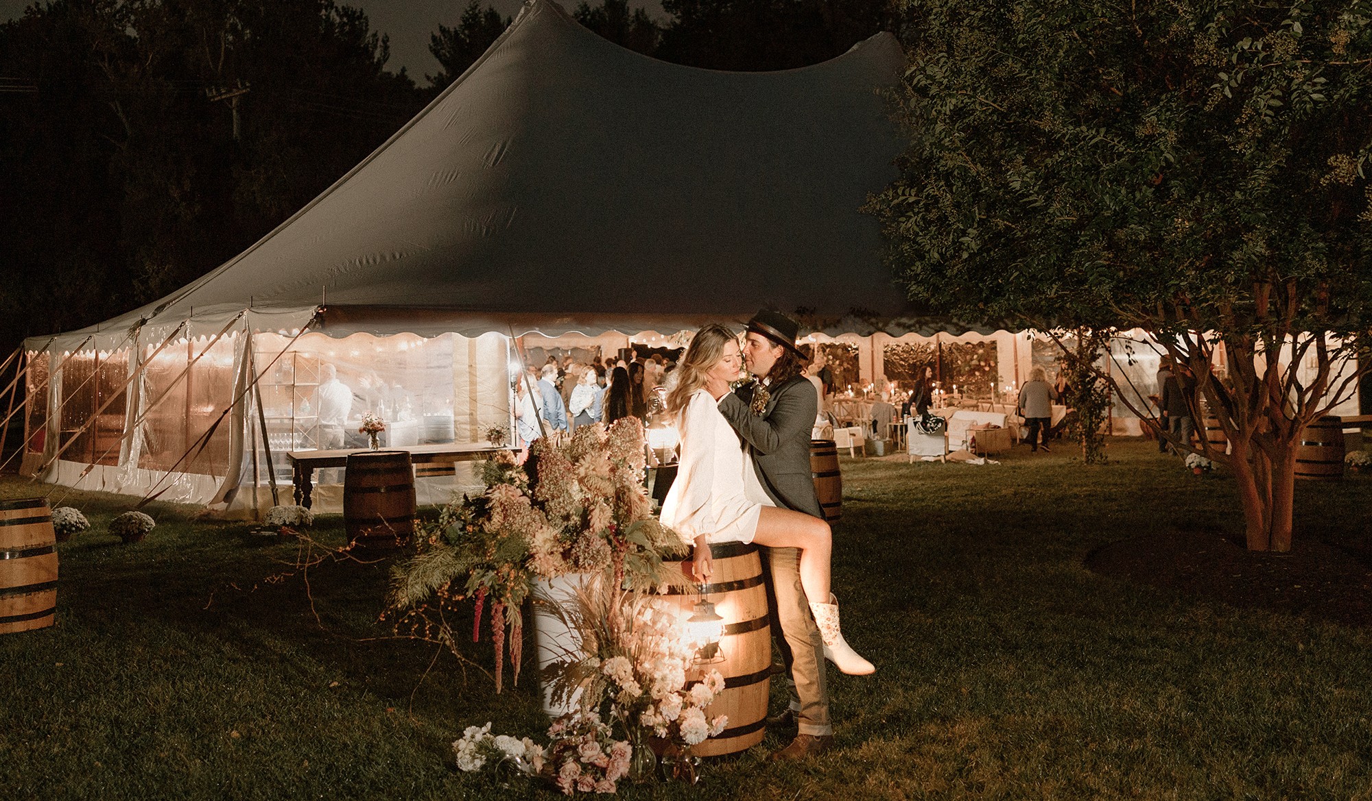 People sitting on barrels under a large outdoor tent at night, with trees and string lights in the background.