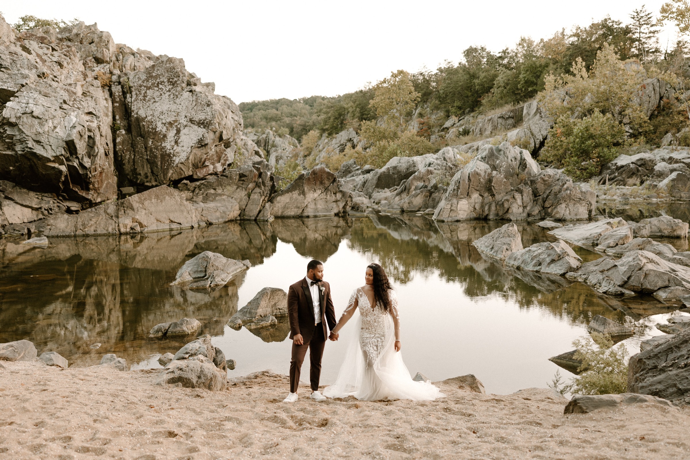 A couple in wedding attire holding hands on a rocky beach near a calm body of water, surrounded by rocks and trees.