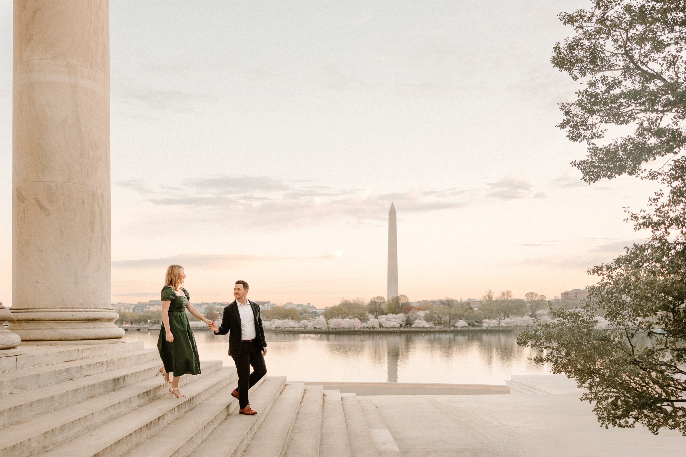 Two people stand on steps near a body of water with the Washington Monument in the background, trees on the right side.