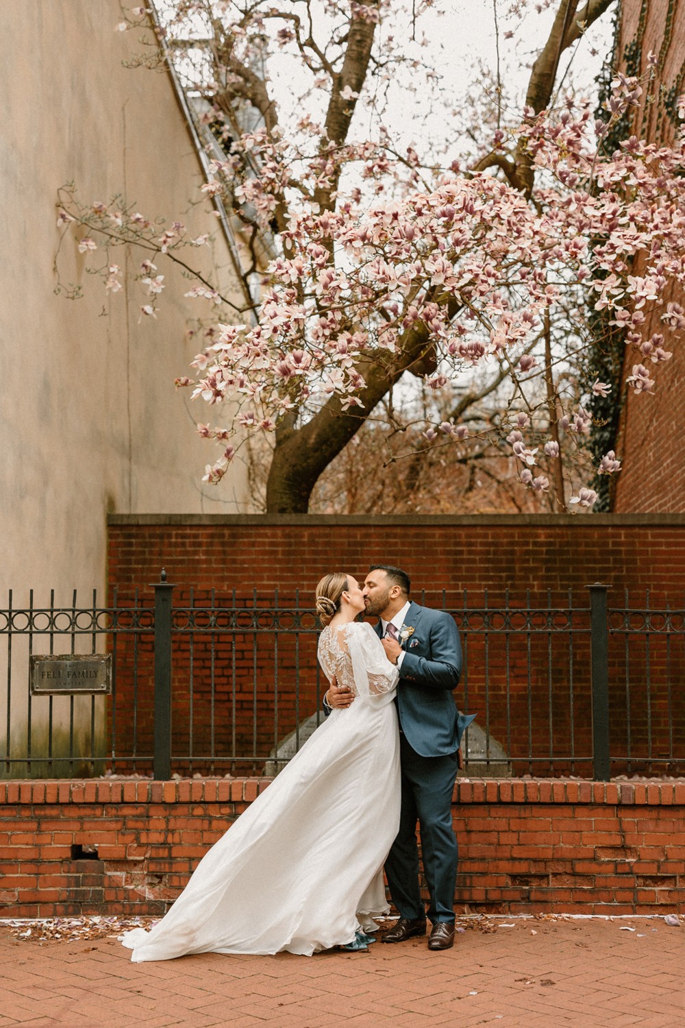 A couple in wedding attire kissing in front of a brick wall with a cherry blossom tree overhead.