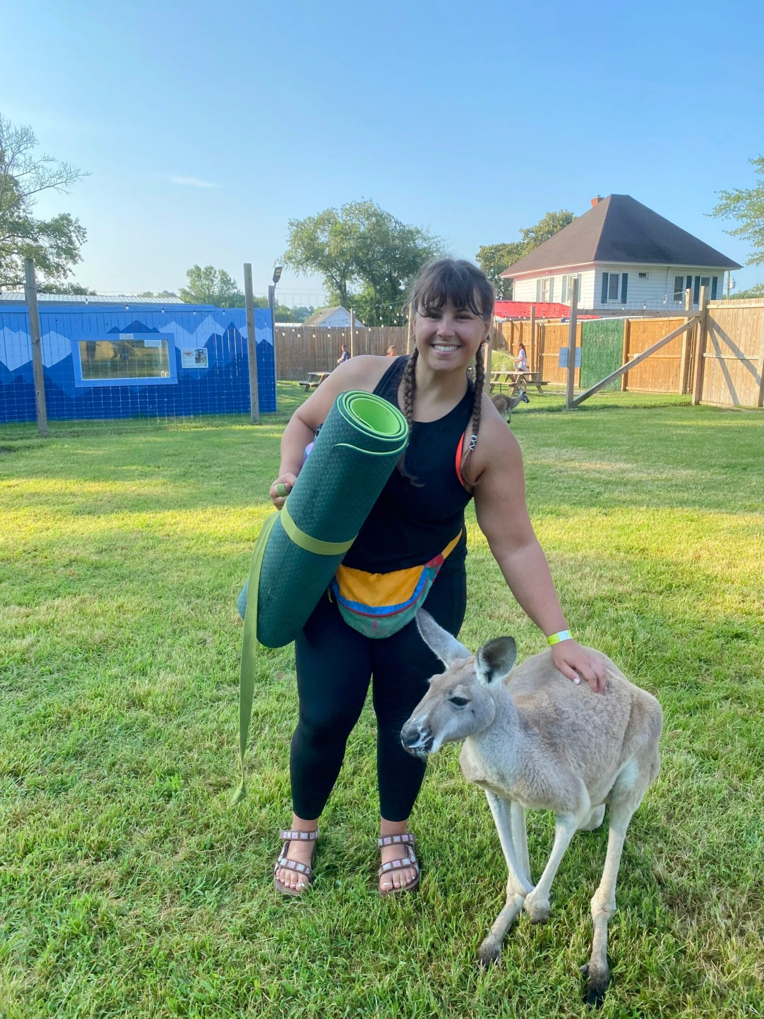 Woman standing outdoors on grass, holding a yoga mat, petting a small goat, with trees and buildings in background.