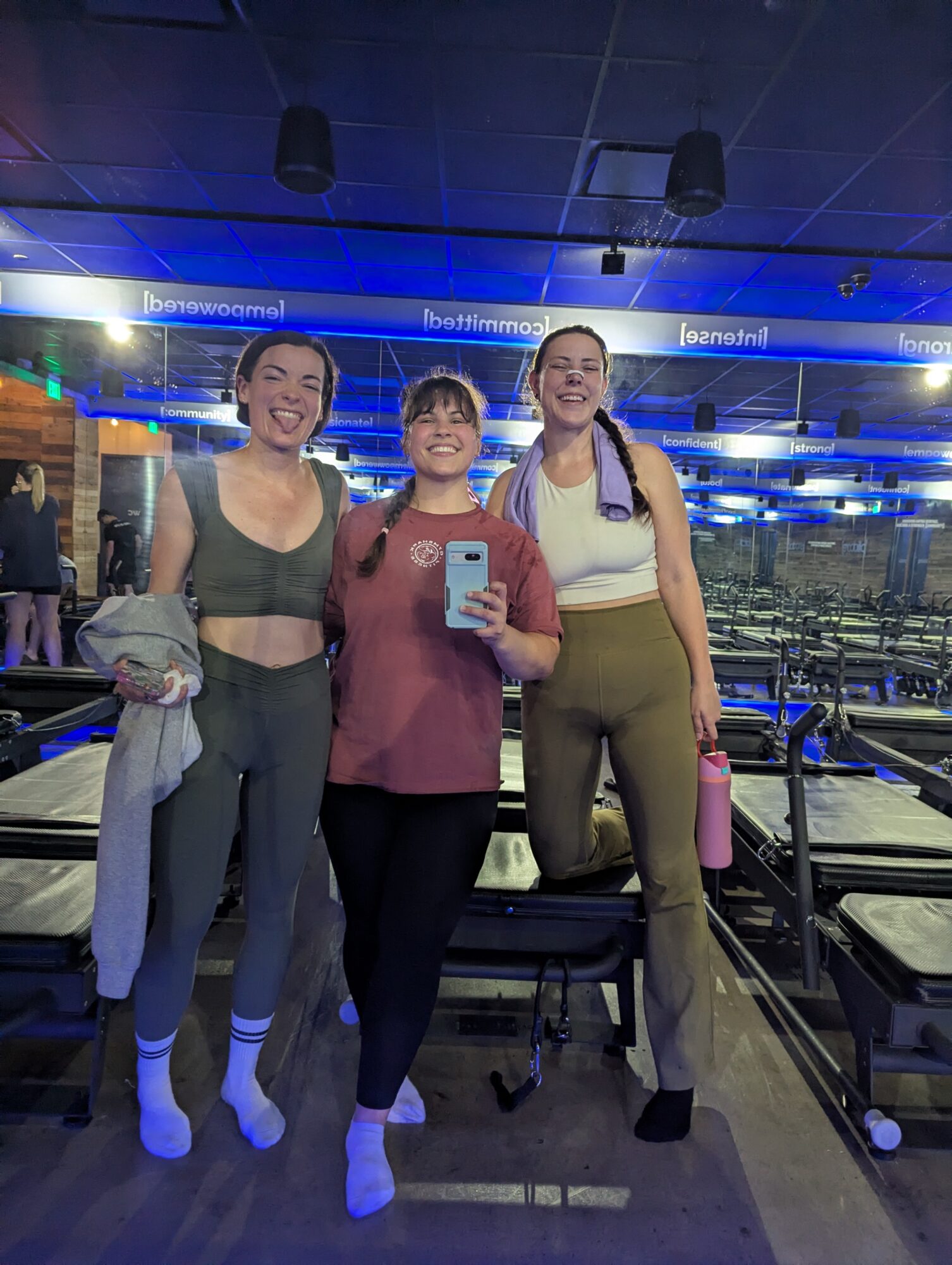 Three women taking a mirror selfie in a gym, smiling, with exercise equipment in the background.