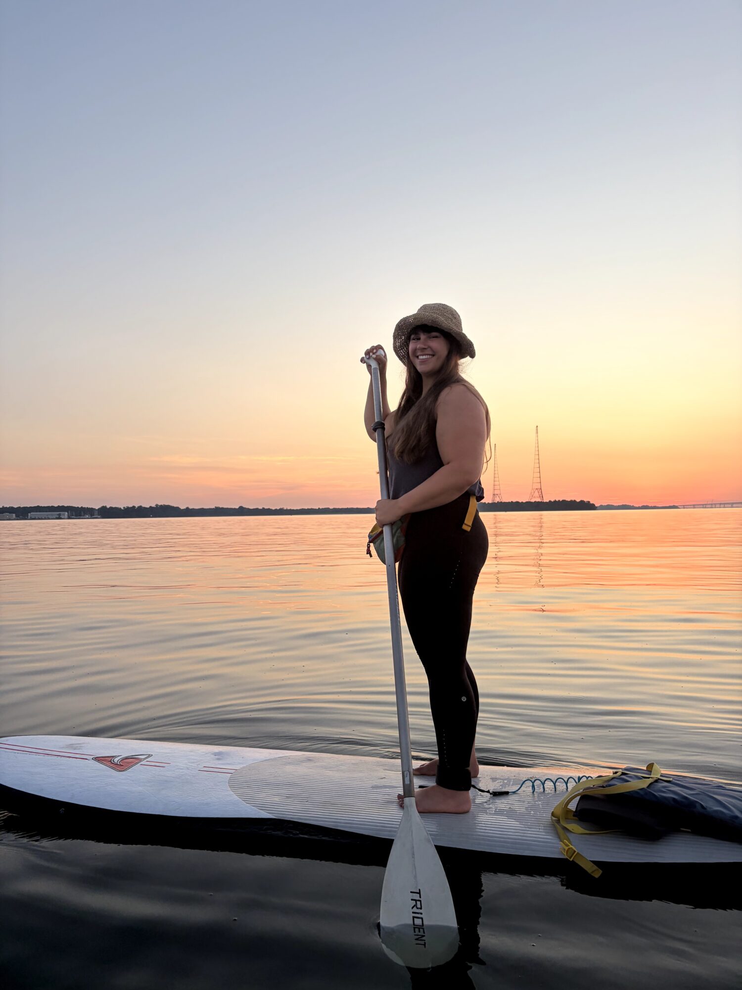 Woman standing on paddleboard on water during sunset, holding paddle, wearing hat and sunglasses.