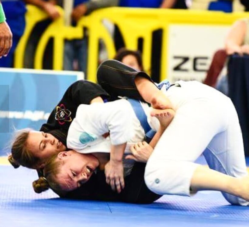 Two women competing in Brazilian Jiu-Jitsu on a blue mat, one in a white gi and the other in a black gi, grappling intensely.