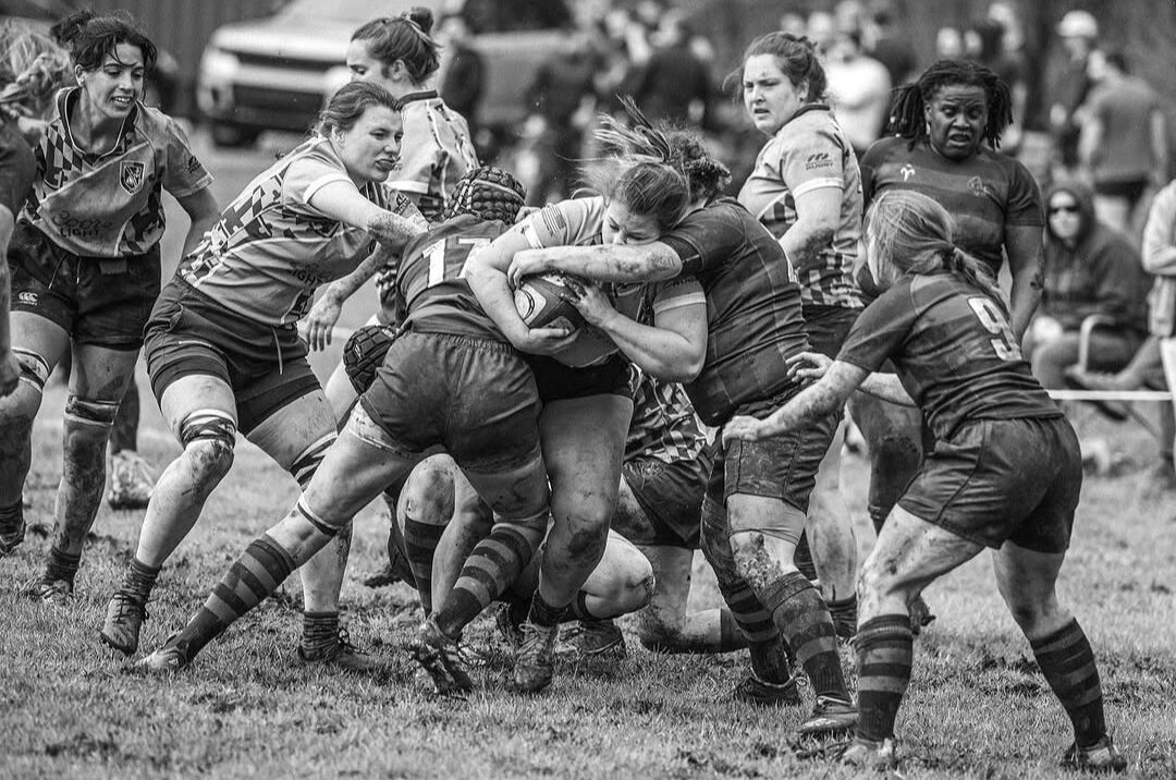 Rugby match with players engaged in a tackle on a grassy field, spectators in the background, black and white image.