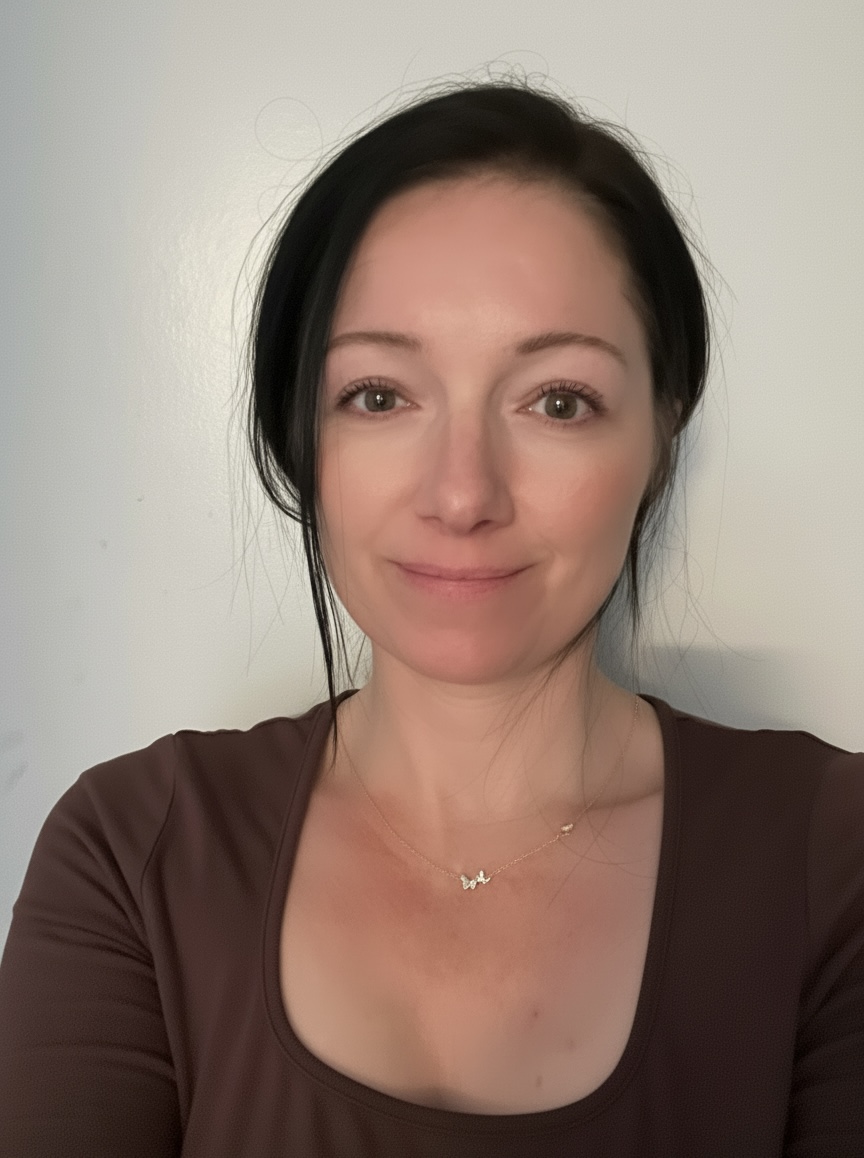 Woman with dark hair and light skin, wearing a brown top and a necklace, smiling softly against a plain background.