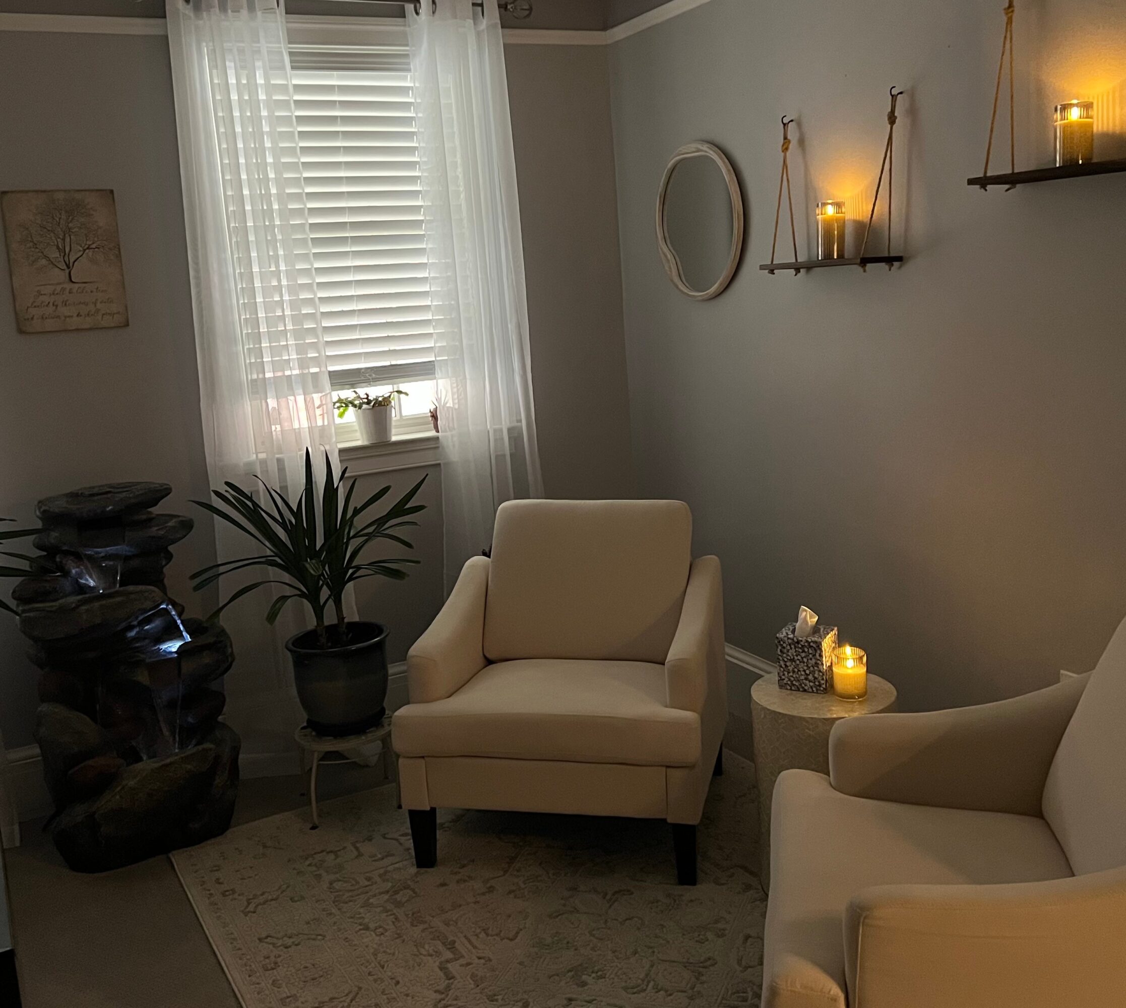 Cozy living room corner with armchair, plant, window with curtains, wall decor, and candles on shelves and table.