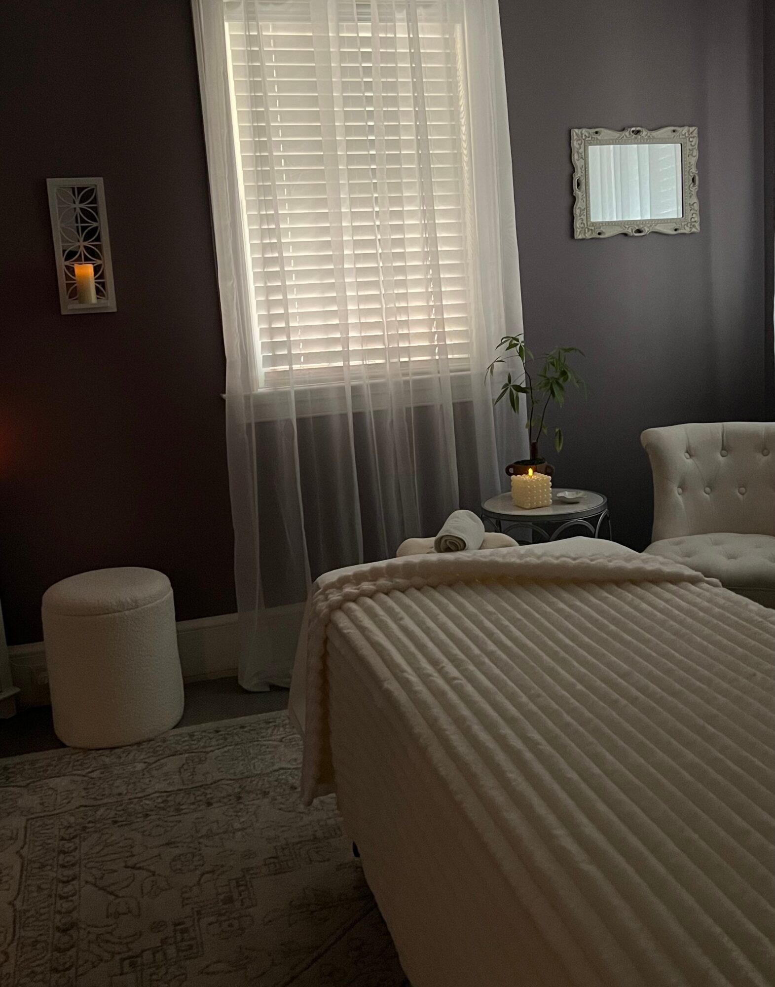 Bedroom with window, sheer curtains, white bedspread, and a small side table with a plant and candle, cream-colored armchair, and wall decor.