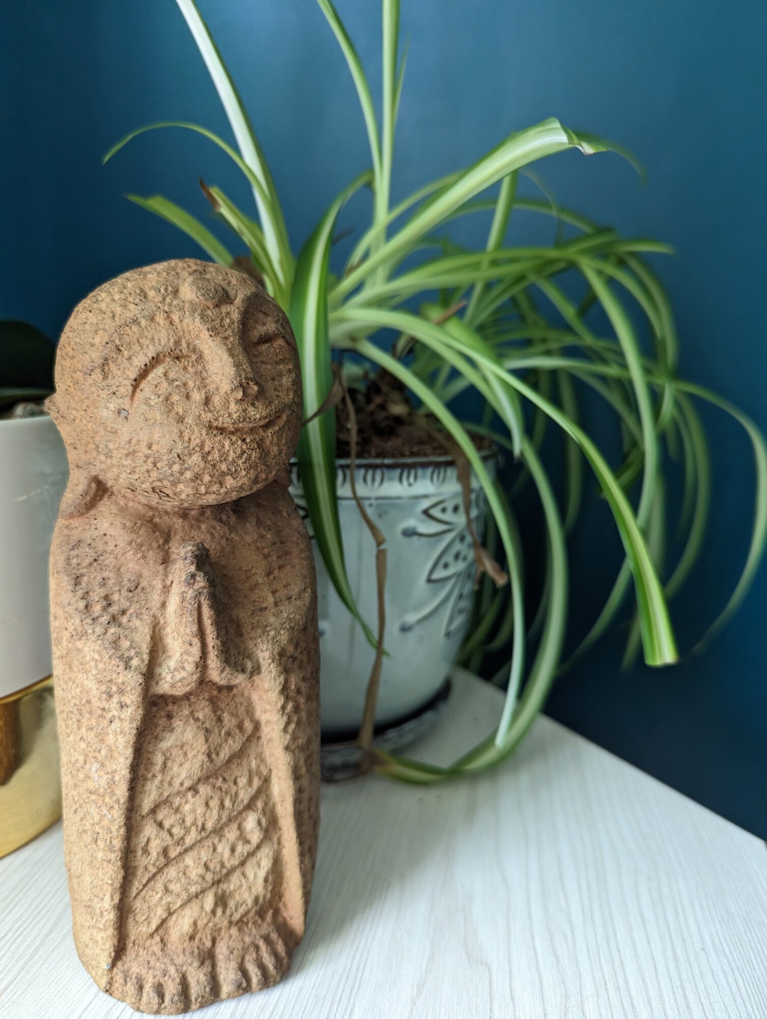 Stone sculpture of a smiling figure with a rounded head, standing next to a potted plant with long, green leaves, on a white surface.