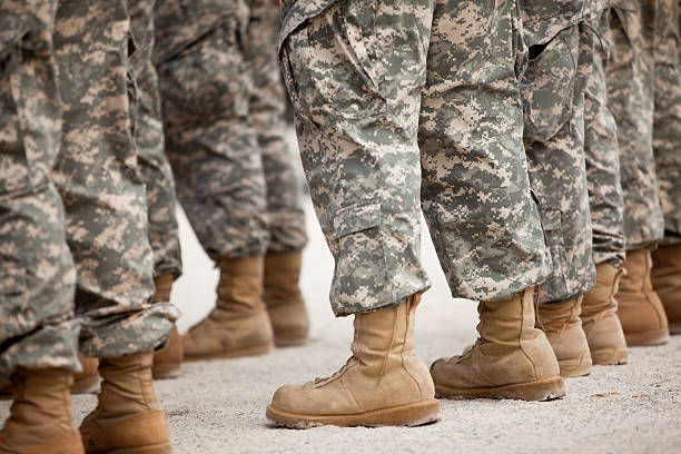 Line of soldiers in camouflage uniforms and tan boots standing on ground, viewed from the side.