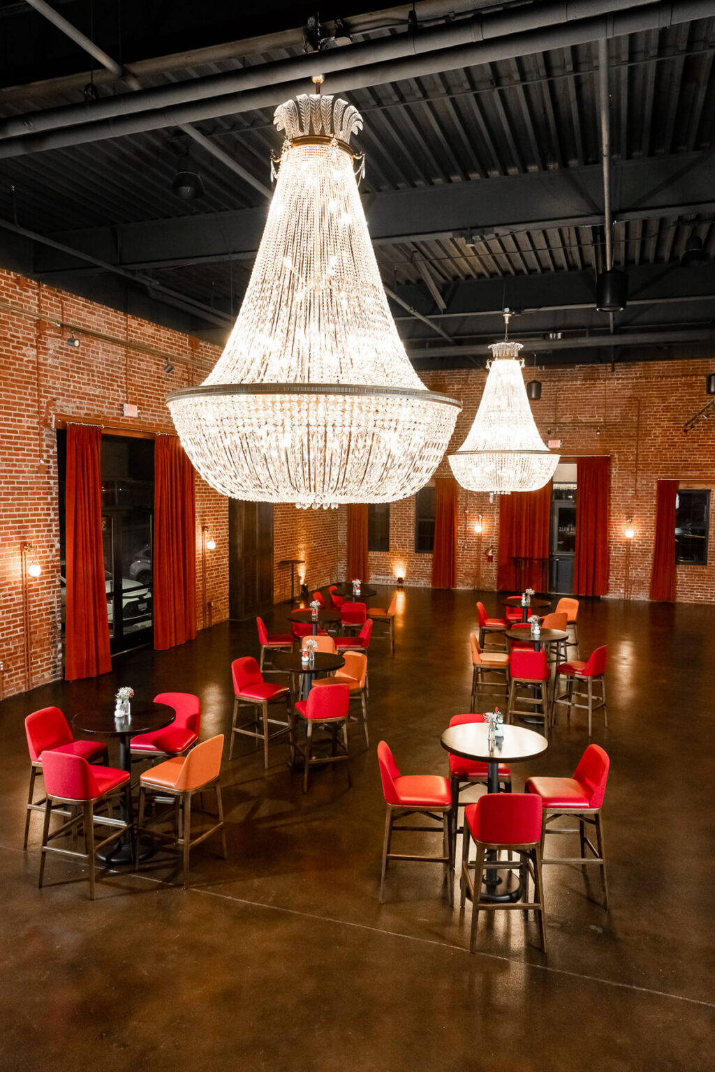 Elegant dining area with red chairs, round tables, and two large chandeliers hanging from a dark ceiling.