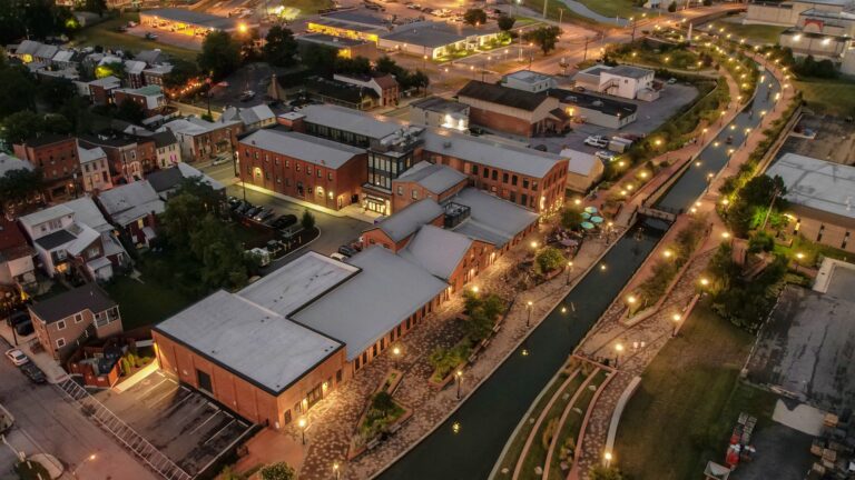 Aerial view of a cityscape at dusk with illuminated streets, buildings, and a canal running through the area.