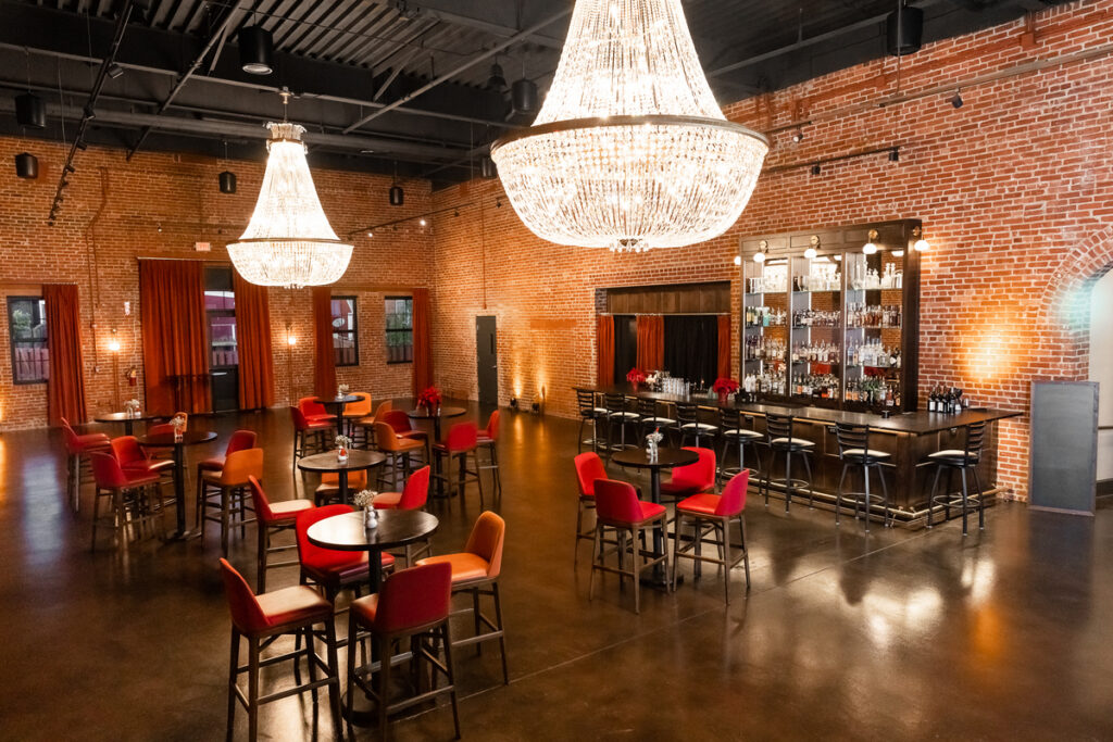 Elegant event space with chandeliers, brick walls, and tables with red and orange chairs, set for a gathering.
