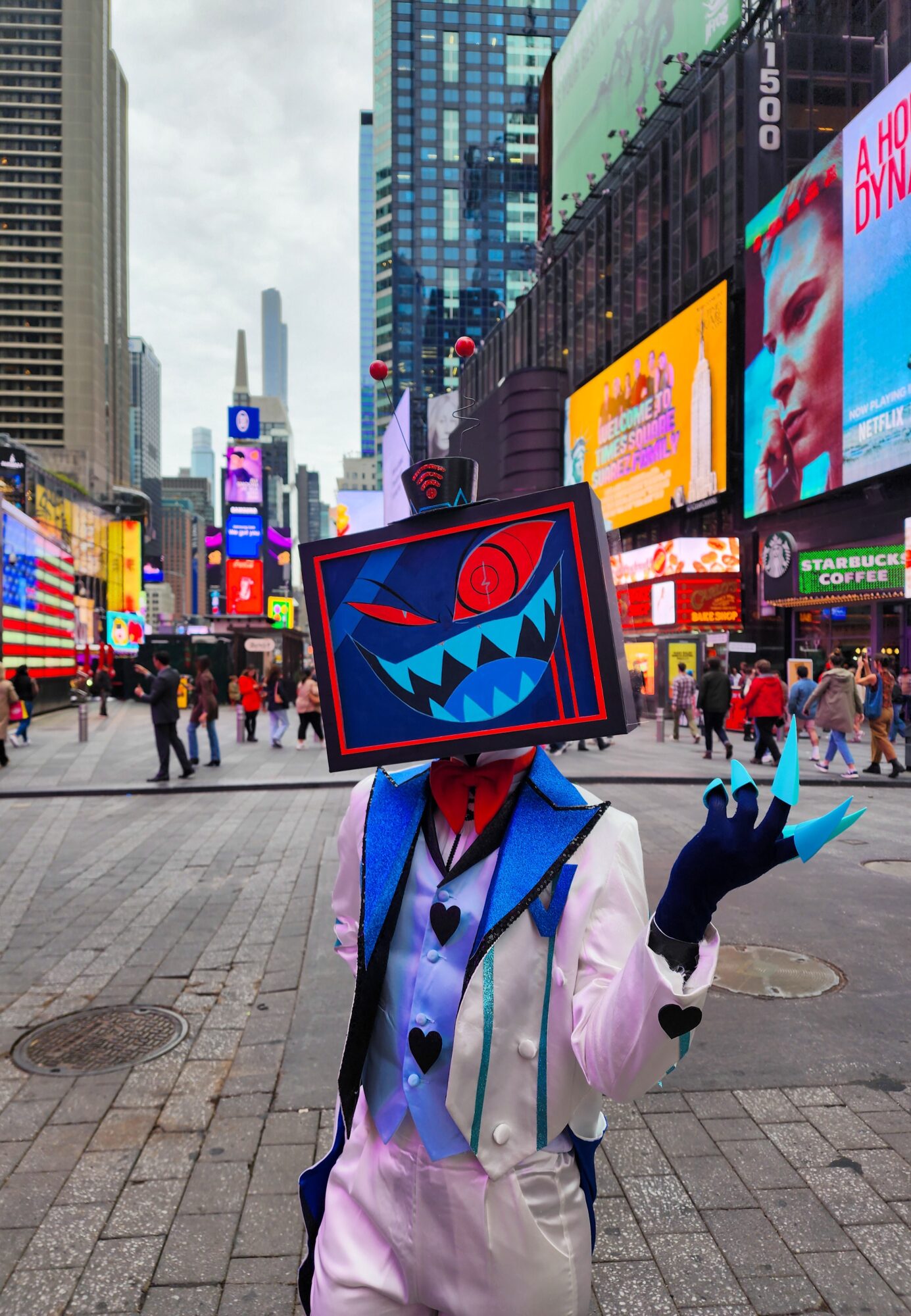 Person wearing a costume with a TV screen head displaying a cartoon face, standing in a busy city square with digital billboards.
