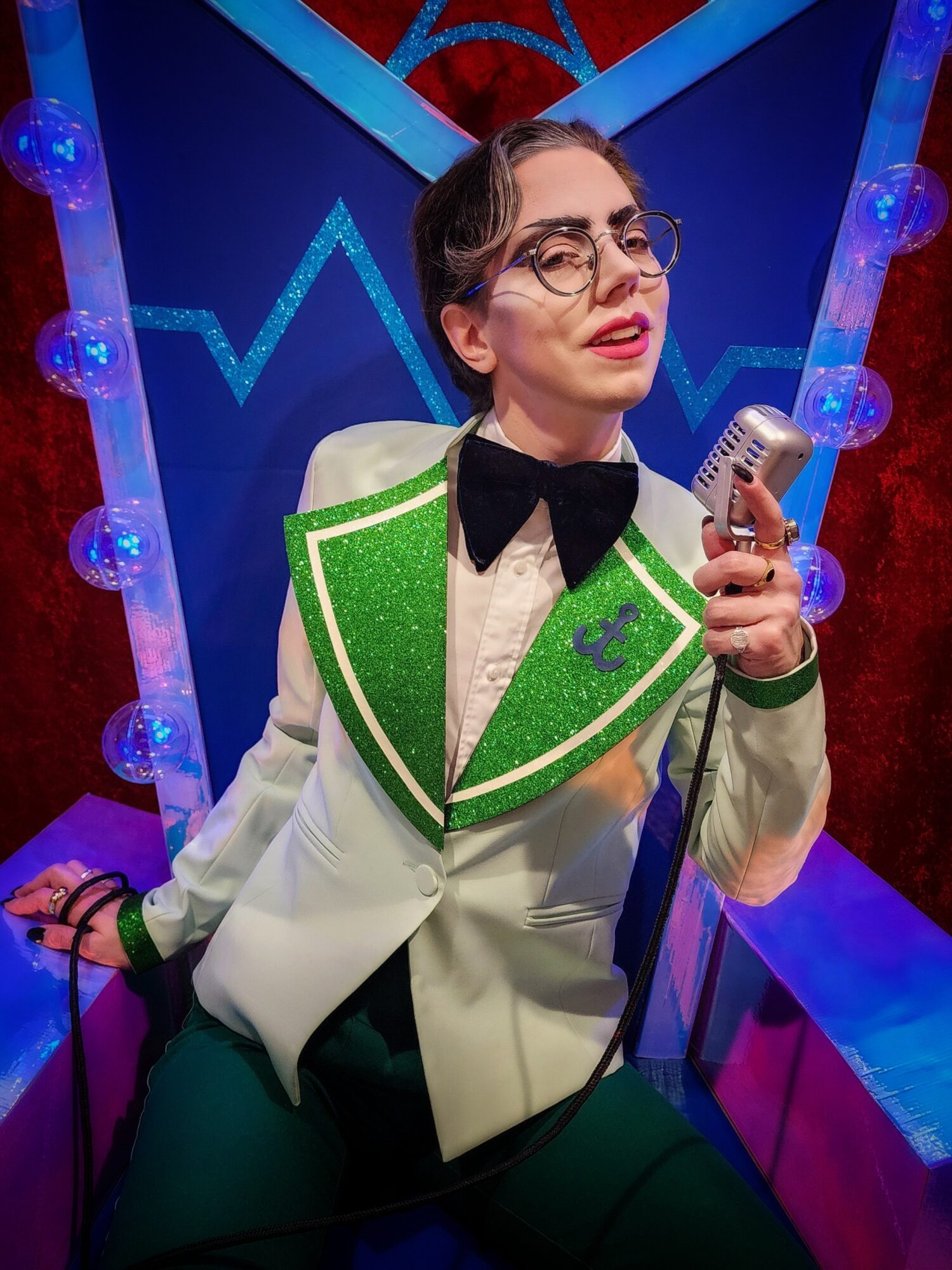 Woman with glasses and dark hair in a white and green costume holding a vintage microphone, standing in a colorful set.