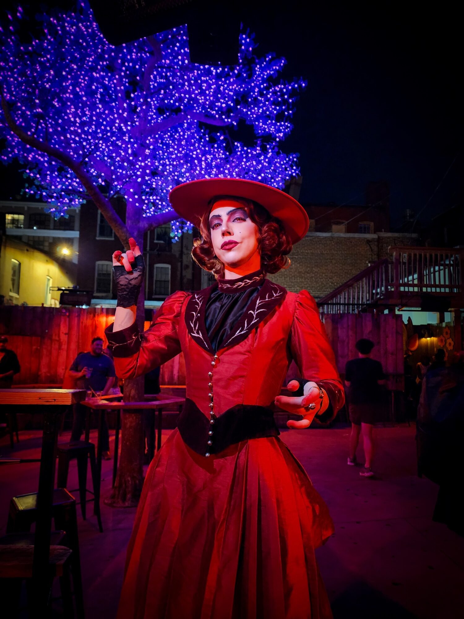 Woman in red dress and hat standing outdoors at night with illuminated blue tree in background.
