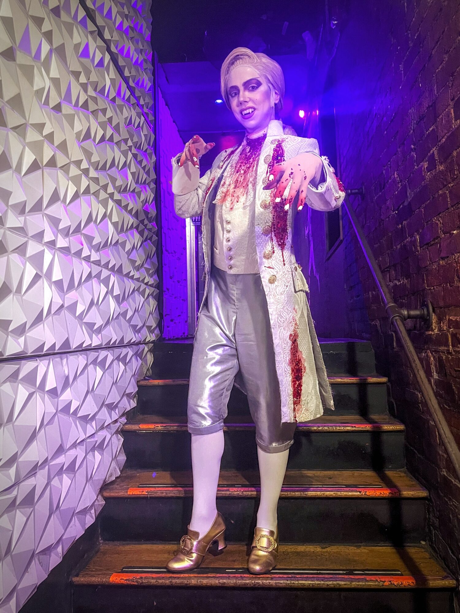 Person dressed in historical costume with white coat, knee-length pants, and gold shoes, standing on stairs in a dimly lit setting.
