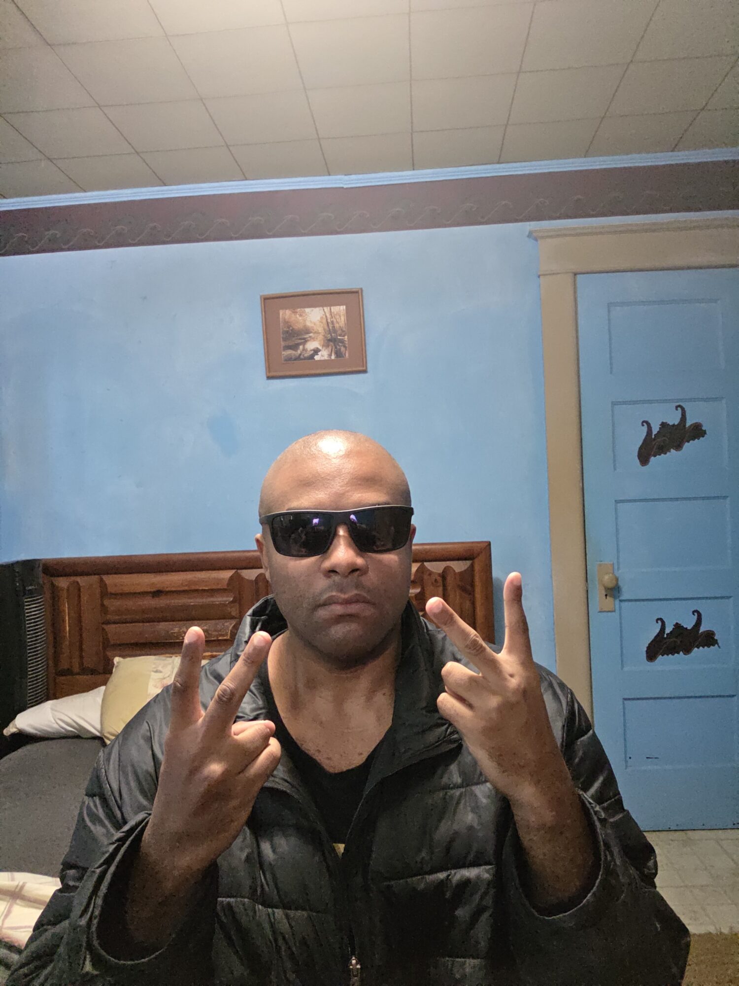 Man with sunglasses making peace signs with both hands indoors, blue door and wall behind him.