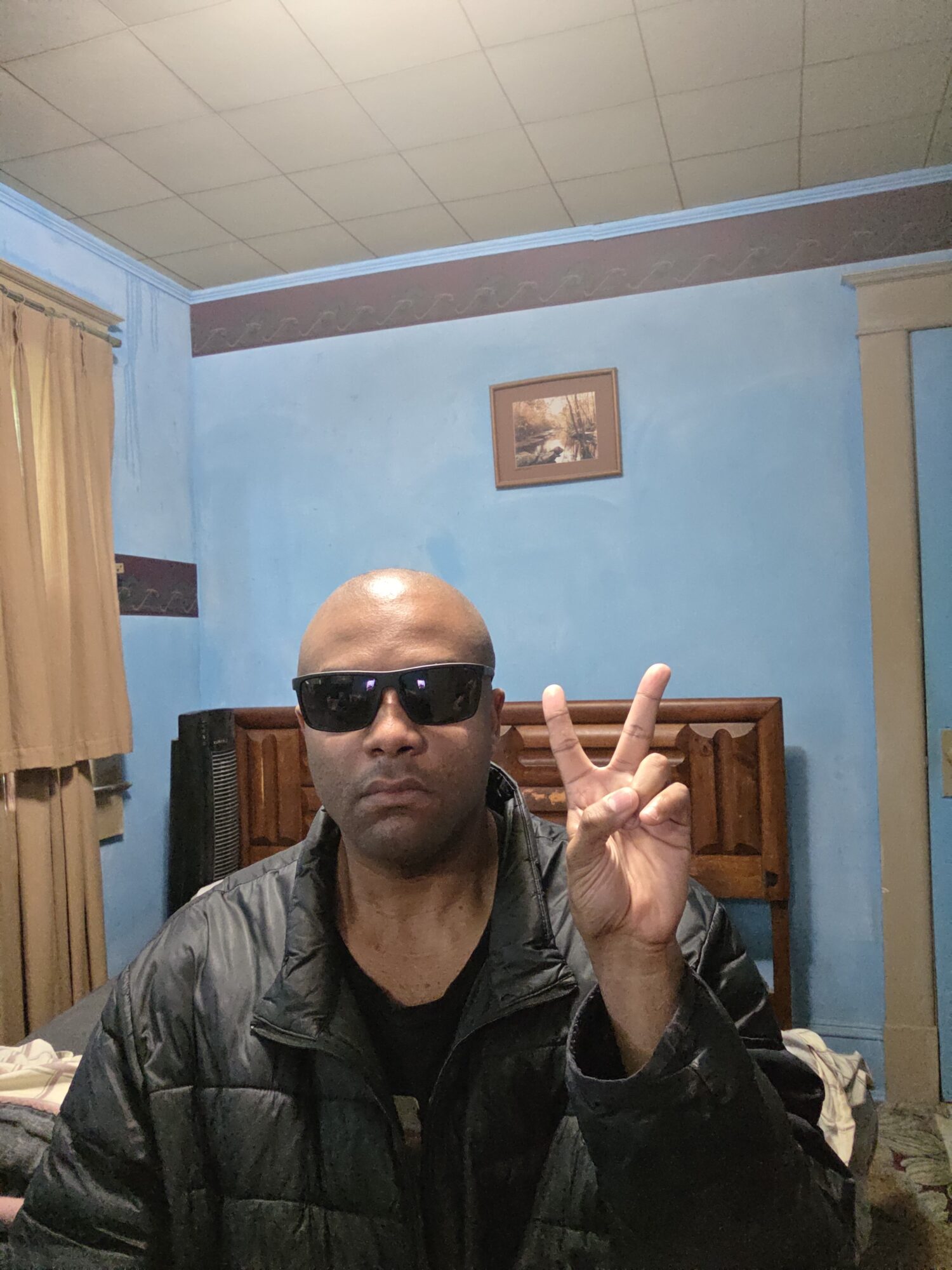 Person with sunglasses making a peace sign in a room with blue walls and a wooden headboard.