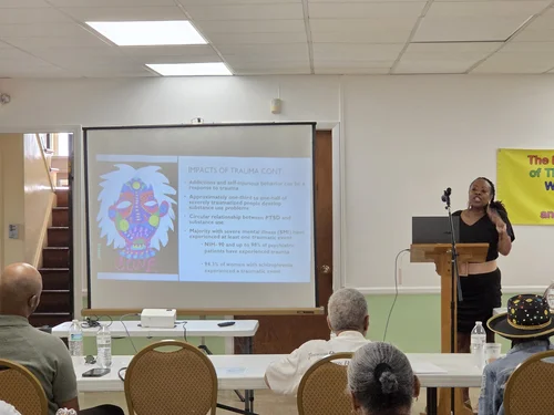 A woman stands at a podium giving a presentation in a room with seated audience members, a large screen displaying a colorful graphic, and a yellow banner on the wall.