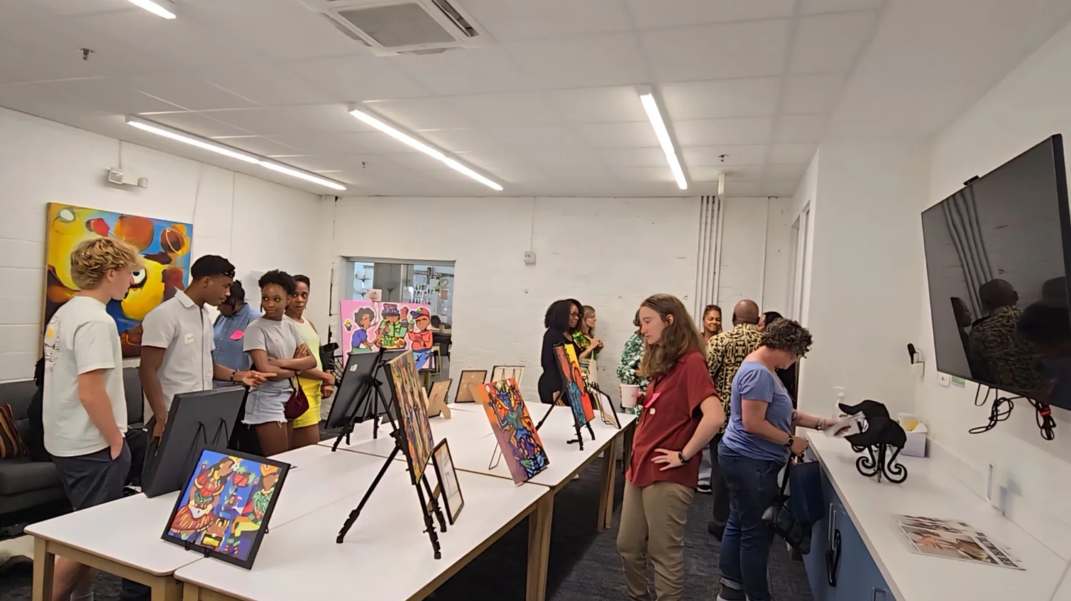 Group of people in a room with artwork and display tables, some looking at art and others talking.