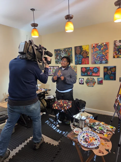 Person being interviewed in art gallery, colorful paintings on wall, table with art supplies, camera operator filming.