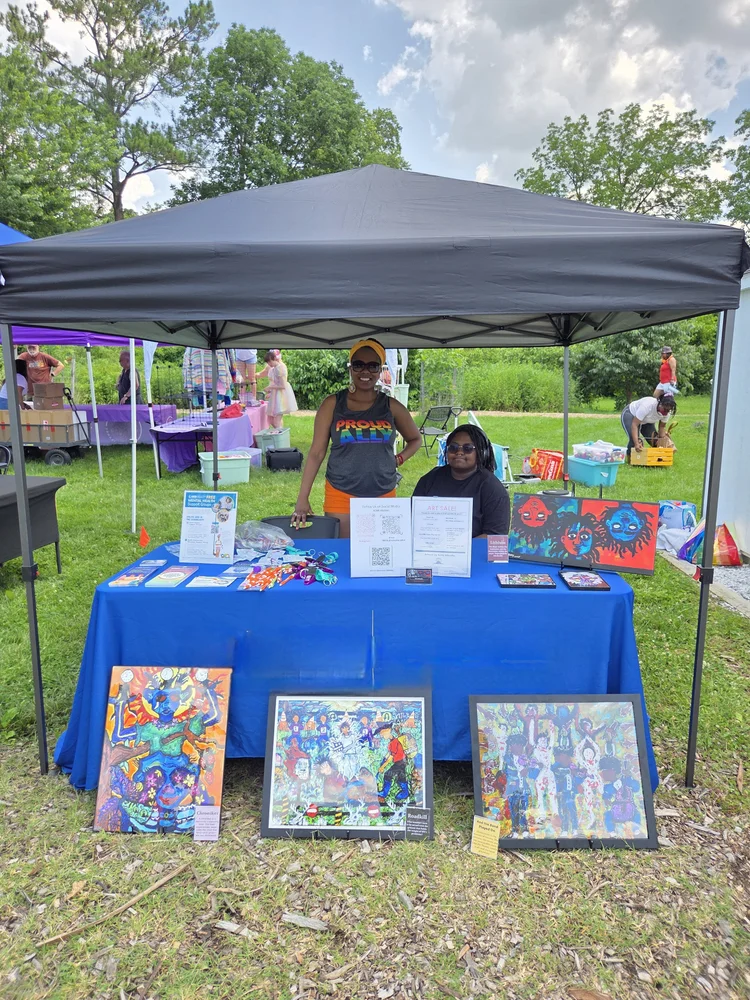 Two people stand behind a table with colorful artwork under a black canopy in a grassy outdoor area.