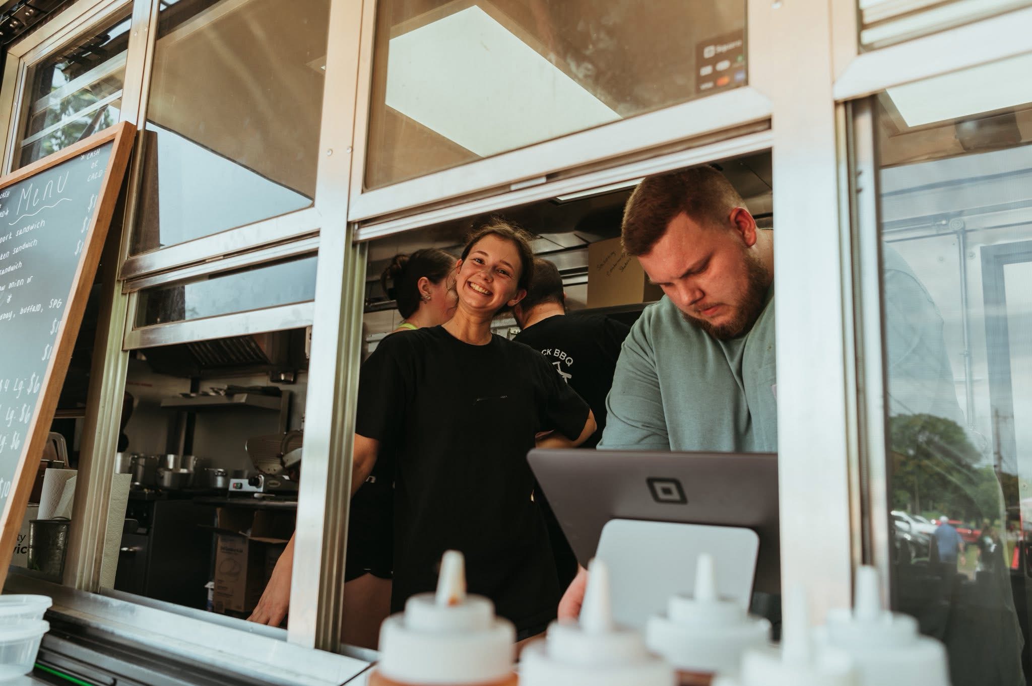 People inside a food stand, one person working on a laptop, others smiling and talking, with condiment bottles in foreground.